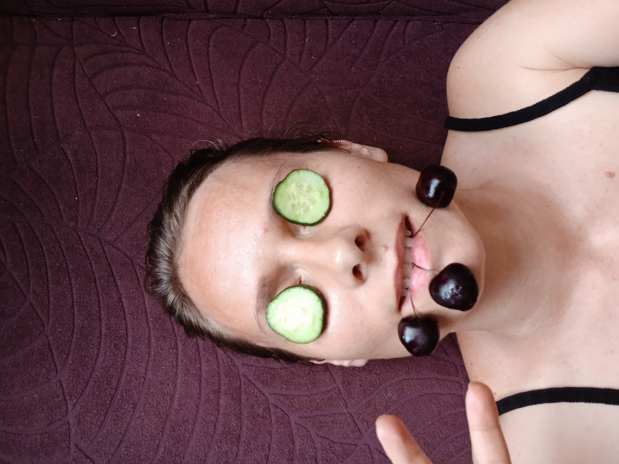 A young Caucasian woman with dark hair, wearing a black tank top, is lying down on a dark purple textured surface. Slices of cucumber are placed over her eyes, and two cherries are held between her teeth by their stems. Her left hand is raised and visible in the lower left quadrant of the image, with her index finger extended. The scene appears to be an indoor setting, possibly a bedroom, given the textured background which resembles a blanket or upholstered surface. There is no indication of the city of Yekaterinburg, Russia, within the image.