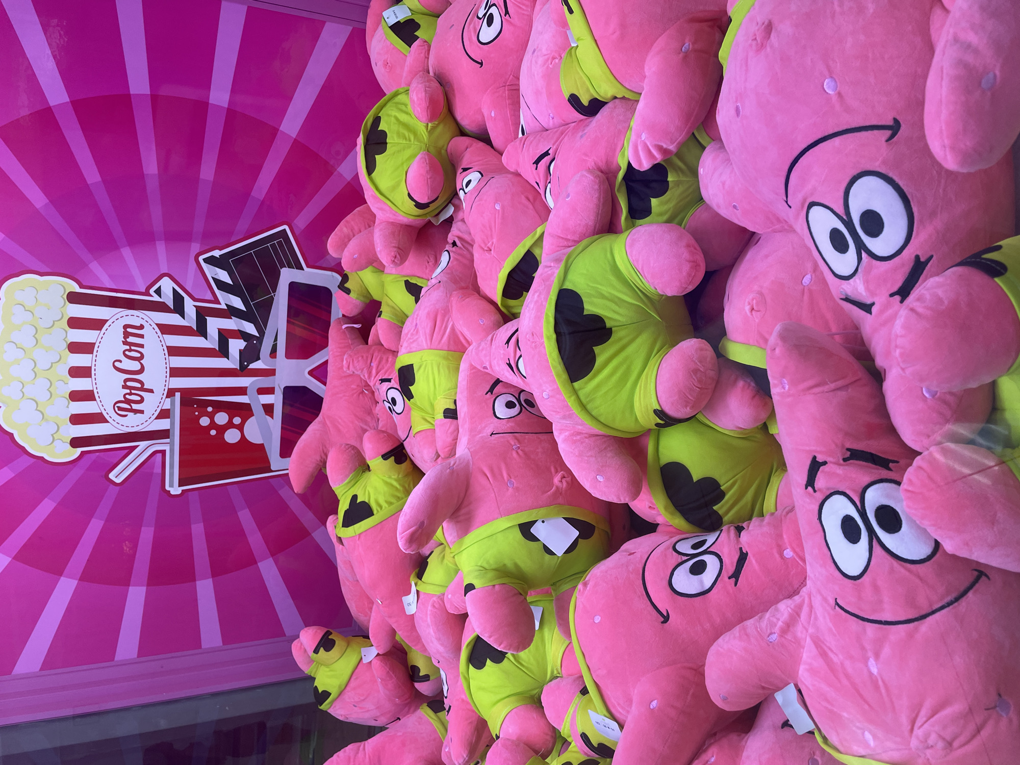 A pile of large, pink plush toys resembling Patrick Star from SpongeBob SquarePants is crammed into a claw machine. The toys are all wearing lime green shorts. The background of the claw machine is pink with a radial stripe pattern and features graphics of popcorn, a soda cup, 3D glasses, and a clapperboard, suggesting a movie theme. The image appears to be taken in a commercial establishment, possibly an arcade or entertainment center, but no specific location in Sochi, Russia is identifiable from the visual information provided.