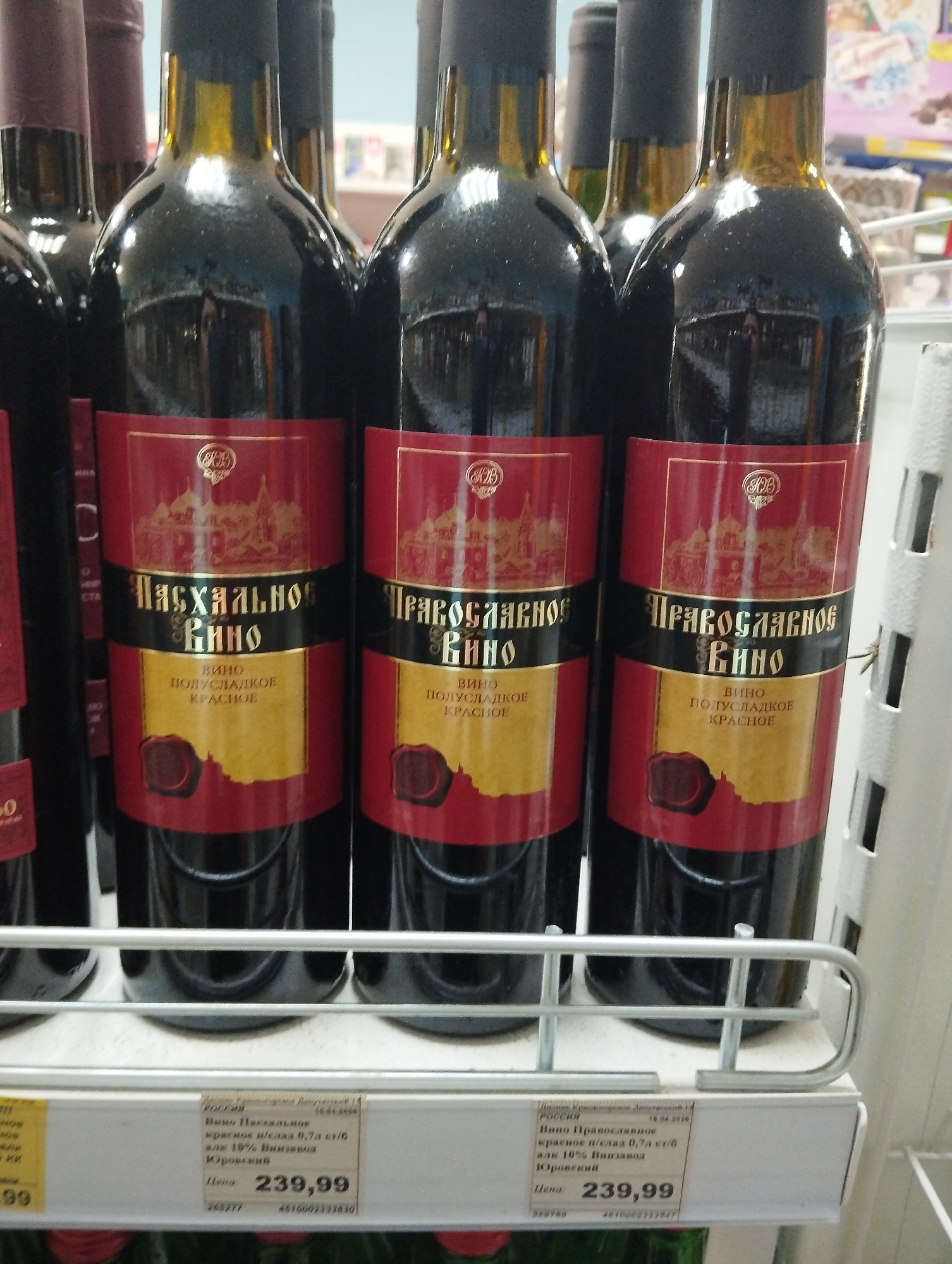 The image displays a close-up of several dark glass wine bottles arranged on a metal shelf in what appears to be a store or supermarket. The bottles are uniformly dark, reflecting the surrounding environment slightly.
The central focus is on three bottles in the foreground, each featuring a distinctive red and gold label. The labels on all bottles read "ВИНО ПОЛУСЛАДКОЕ КРАСНОЕ" (WINE SEMI-SWEET RED). The bottle on the left is labeled "Пасхальное Вино" (Paschal Wine), while the two bottles on the right are labeled "Православное Вино" (Orthodox Wine). All labels share a gold design of a building, possibly a church or monastery, and a wax seal graphic at the bottom of the gold section, along with a monogram at the top.
Below the bottles, two price tags are visible. Both tags indicate "Цена: 239,99" (Price: 239.99) and state "РОССИЯ" (RUSSIA). The left tag corresponds to the "Paschal Wine," describing it as "красное п/слад 0,7л ст/б алк 10% Винзавод Юровский" (red semi-sweet 0.7L glass bottle alc 10% Yurovsky Winery), with a date of "16.04.2020". The right tag describes the "Orthodox Wine" with identical specifications ("красное п/слад 0,7л ст/б алк 10% Винзавод Юровский") and a date of "16.04.2026". The background is out of focus, showing glimpses of other shelves and products. The setting is a retail environment in Krasnogorskoye, Russia.