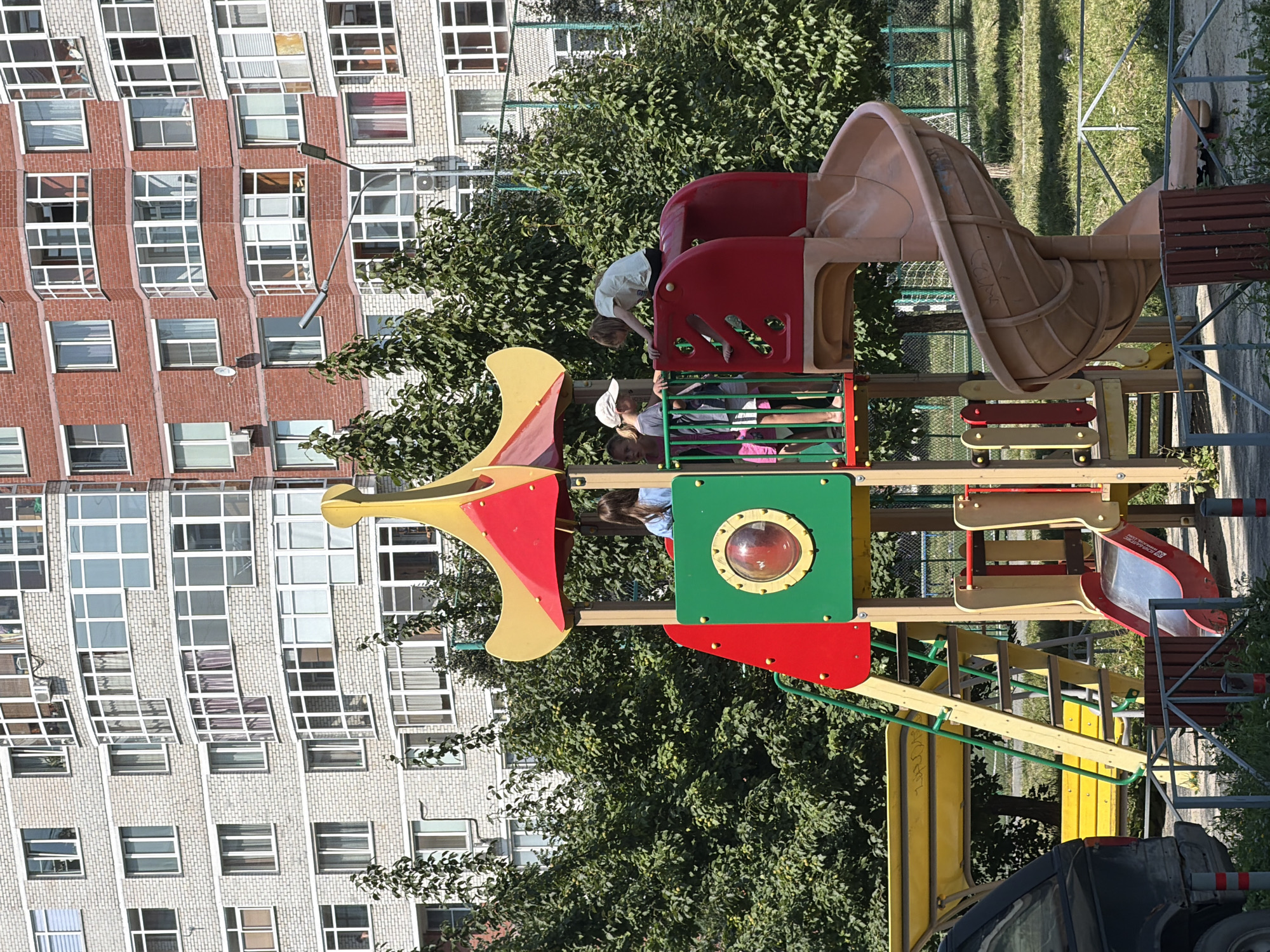 In Yekaterinburg, Russia, an outdoor playground is pictured on a sunny day. The playground features a multi-level structure with slides, a climbing ladder, and a swing set. Four children are visible: three girls on the playground structure and one boy climbing on it. The children are engaged in play. In the background, tall residential buildings with many windows are visible. Trees with green foliage surround the playground. A black vehicle is parked to the left of the frame. The ground is covered in sand and grass.