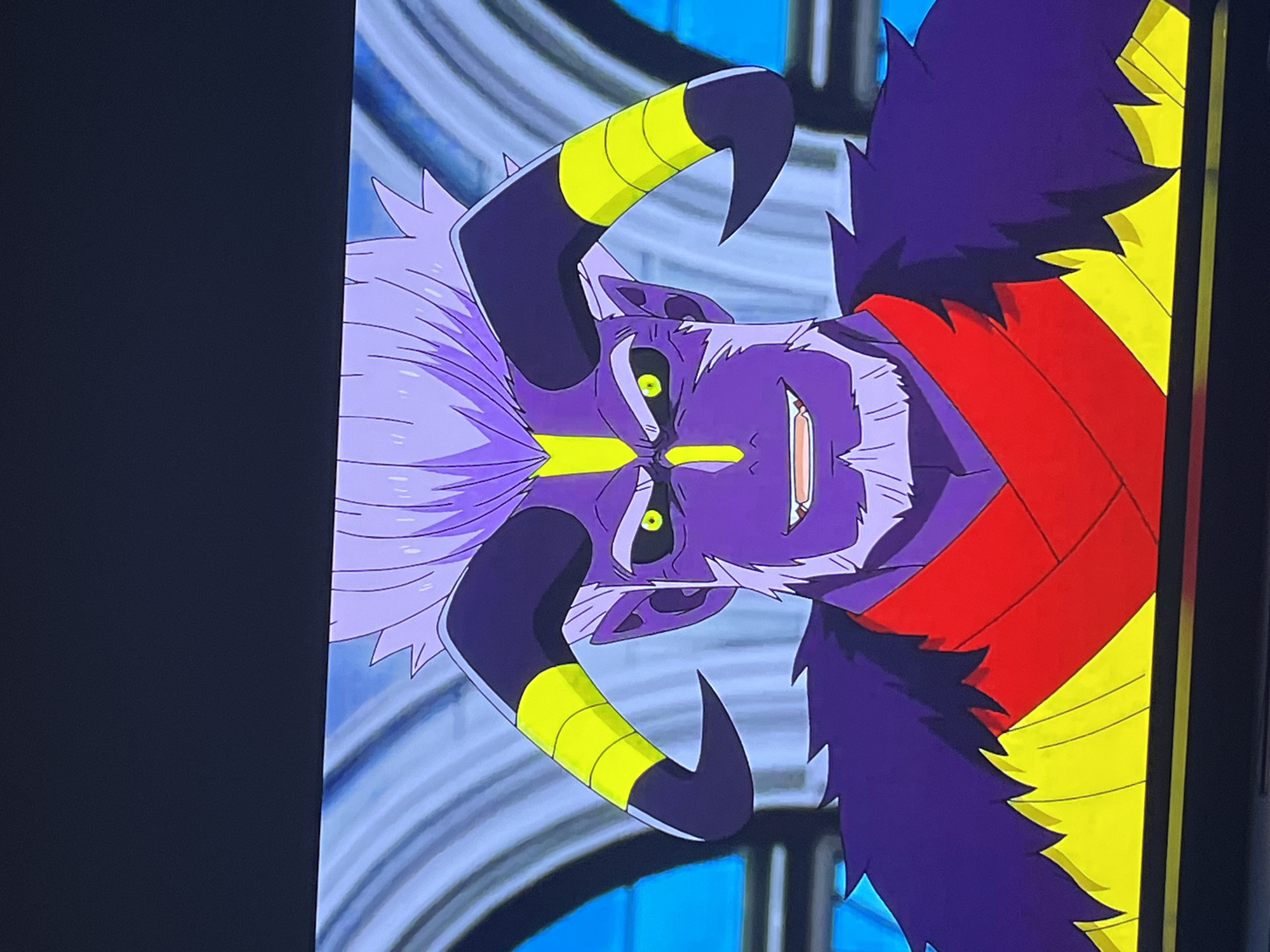 The image is a close-up, eye-level, head-and-shoulders shot of an animated character, appearing to be a demon or a similar mythical being, against a blurred, light-blue and white background that suggests an interior with architectural elements like columns or arches. The character has purple skin, a spiky, light-purple hairstyle, and glowing green eyes with thin, black pupils. Two large, black, curved horns with yellow bands emerge from its head. It has a goatee and a prominent nose and mouth with visible teeth. The character is wearing a dark purple, fur-like garment around its neck and shoulders, over a red garment with yellow trim at the bottom. There is no discernible location in the city of Pilsen, Czechia; the scene is entirely within an animated context.