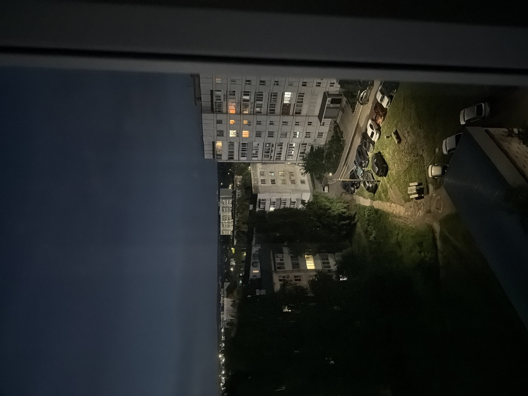 This is a night-time, high-angle, medium shot of a residential area in Chaikovsky, Russia. The sky is dark and overcast, with a hint of light on the horizon. In the foreground, the edge of a window frame is visible on the right side of the image. The mid-ground features several multi-story apartment buildings, some with lights on in the windows, indicating occupancy. Several cars are parked in a lot between the buildings. Trees and patches of grass are also visible. In the distance, more apartment buildings and streetlights can be seen, suggesting a developed urban or suburban environment. The overall impression is one of a quiet, possibly sleeping, neighborhood at night.
