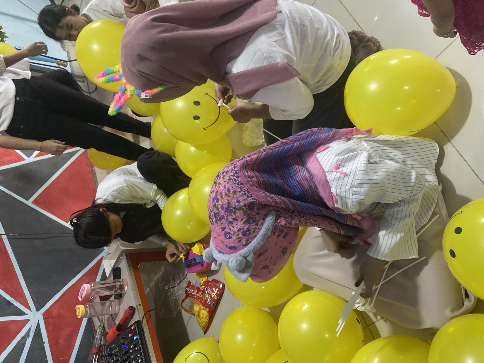 In Tanjung Gading, Indonesia, a group of young people are gathered on the floor, surrounded by yellow balloons, likely preparing for a celebration. The room features a geometric wall with red, black, and light blue patterns. On a table to the left, a mixing board, a red microphone, and two water bottles are visible. One of the individuals, wearing a pink and purple headscarf, is in the process of attaching a balloon to a stick. Another person in a white shirt is also holding a balloon, possibly inflating it, while a third person in a white shirt and black pants stands in the background, partially visible. The floor appears to be tiled, and the overall atmosphere suggests a casual, festive gathering.