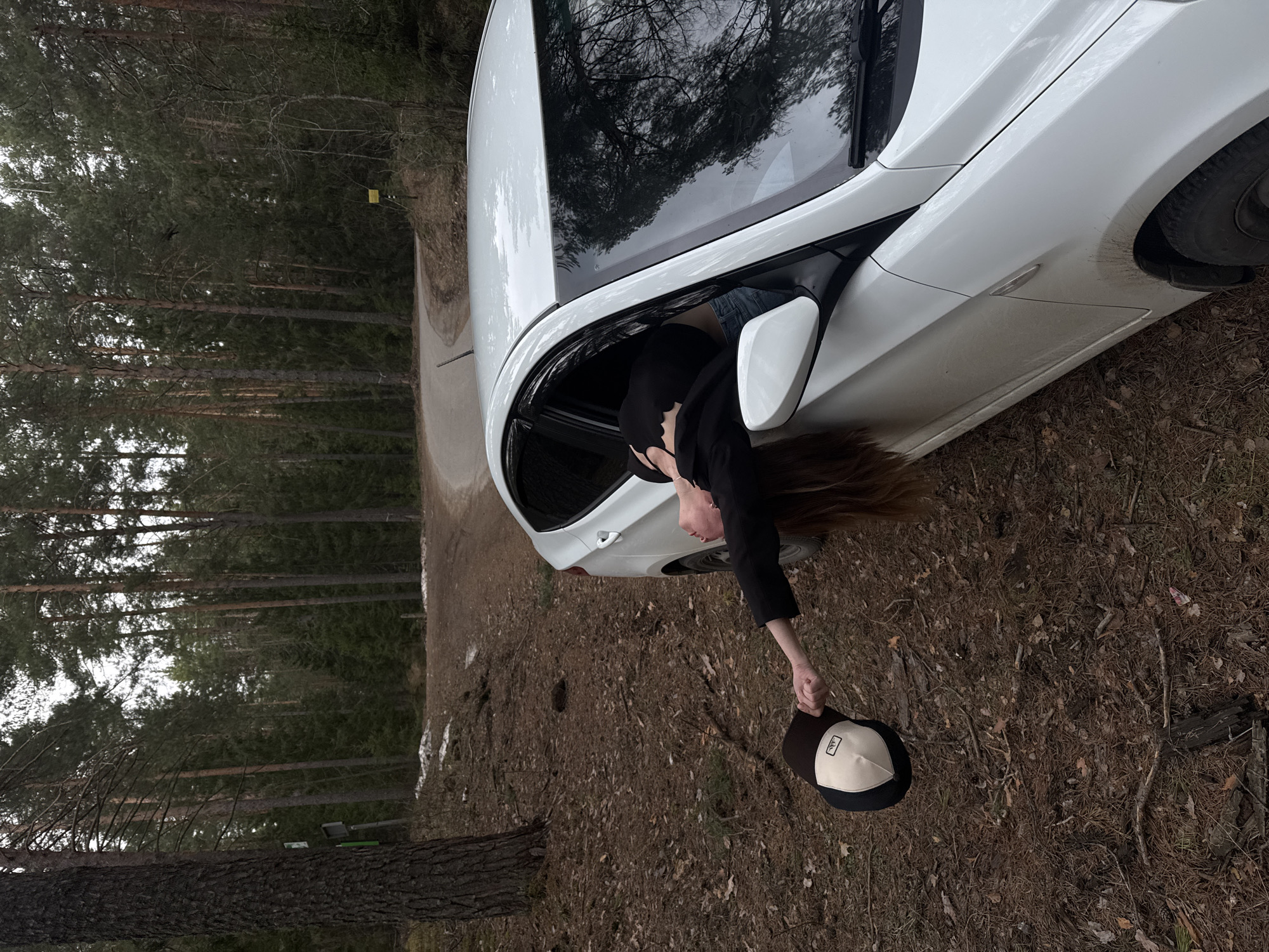 This media file captures a playful moment in a forest environment, likely near Chaikovsky, Russia. The image is taken during the daytime under an overcast sky, as indicated by the diffused light and lack of harsh shadows.
The main subject is a person, appearing to be female, with long brown hair, leaning out of the open passenger-side window of a white sedan. The car, possibly a BMW given its styling, is parked or slowly moving on what appears to be a dirt or gravel road. The person is wearing a dark long-sleeved top and is holding a two-toned baseball cap in their outstretched hand, dangling it towards the ground. The cap has a light-colored front panel with an illegible dark logo (resembling a stylized "AAA") and a dark brim and back. The person's face is partially visible, looking downwards.
The car's white body shows some dirt and mud splatters on its lower panels and wheel arch, suggesting it has traveled on unpaved terrain. The car's windows and roof reflect the surrounding trees, adding depth to the scene.
The setting is a dense forest, characterized by numerous tall coniferous trees, likely pines, with some deciduous trees showing bare branches, possibly indicating early spring or late autumn. The forest floor is covered with a thick layer of pine needles and fallen leaves. A narrow, unpaved road runs through the forest, adjacent to the car. In the background, among the trees, a small, square yellow sign is faintly visible. The overall mood of the image is serene and natural.