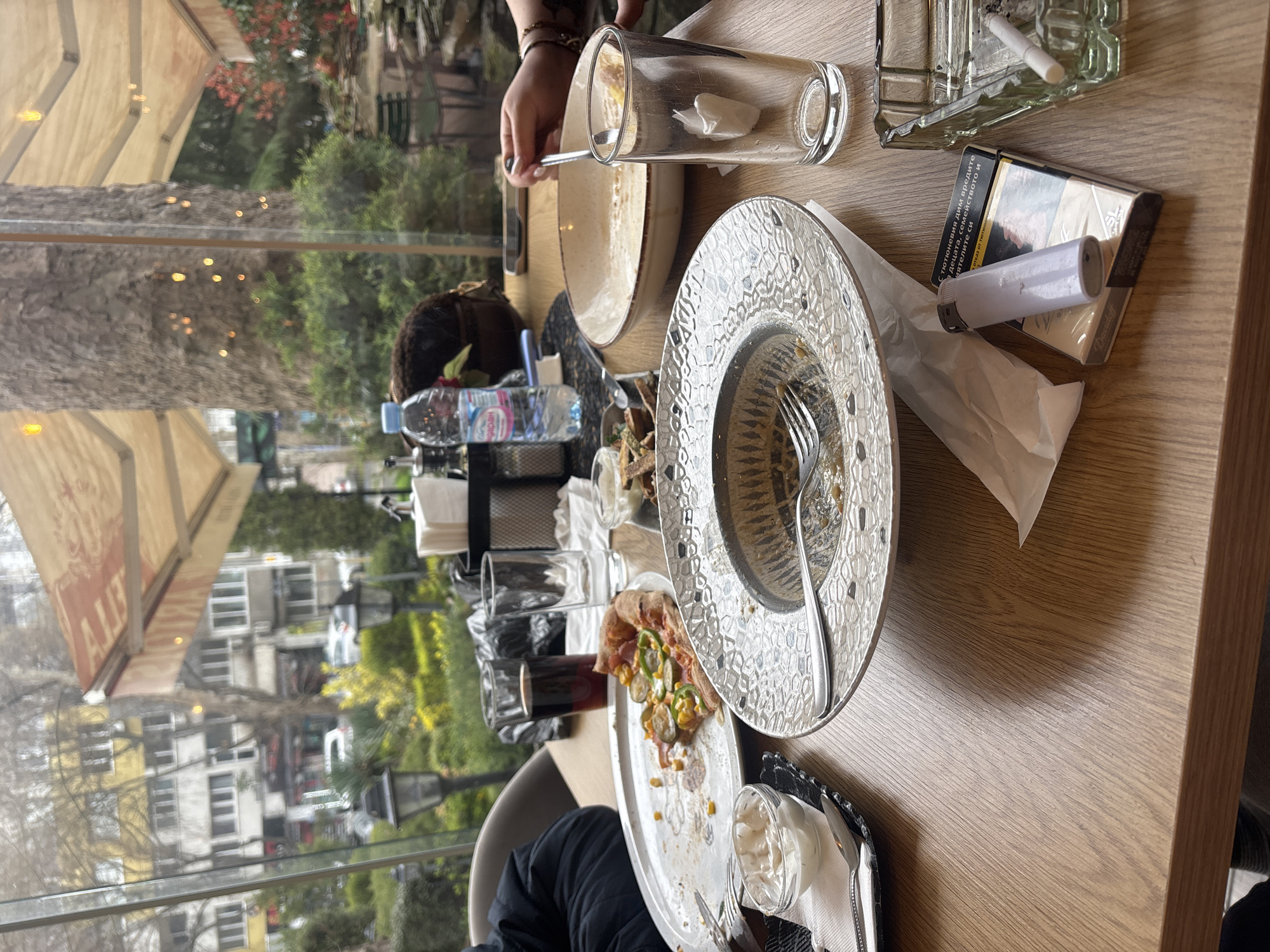 The image depicts a table in a restaurant, likely in Burgas, Bulgaria based on the Cyrillic labeling on the water bottle. The table is cluttered with remnants of a meal: empty plates, some with food scraps, glasses, and silverware. There's a pack of Davidoff cigarettes, a lighter, and an ashtray with a cigarette butt. A person's hand is visible, holding a spoon near a plate. A water bottle is also on the table, along with condiments. Outside the window, buildings and trees are visible, suggesting an urban setting. The overall impression is a post-meal scene with smoking.