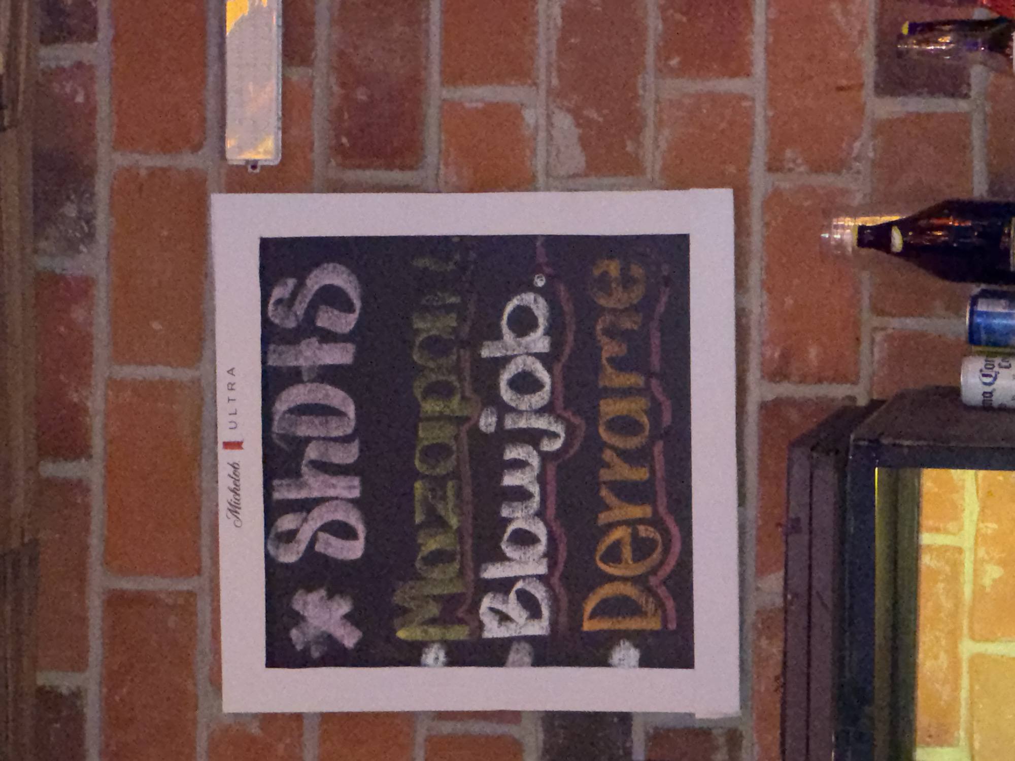 The image depicts an indoor scene in the city of Puebla, Mexico, centered on a chalkboard sign affixed to a reddish-orange brick wall with white mortar. The sign, framed in white, displays "Michelob ULTRA" branding at the top. Below this, "SHOTS" is written in stylized white chalk with an asterisk preceding it. Three shot options are listed: "Mazapan" in yellow-green chalk, "Blowjob." in white chalk, and "Derrame" in orange chalk, each underlined with a wavy line. To the right of the sign, a horizontal, silver-toned reflective strip with a small red end cap is partially visible against the brickwork. Beneath the sign, a dark-colored counter or shelf extends across the lower portion of the frame, illuminated by a warm yellow glow from the left side. On this counter, a dark glass bottle with a clear stopper, another partially obscured bottle, a blue beverage can, and a white can labeled "Corona Cero" are present. No people are visible. The setting suggests a bar or casual establishment.