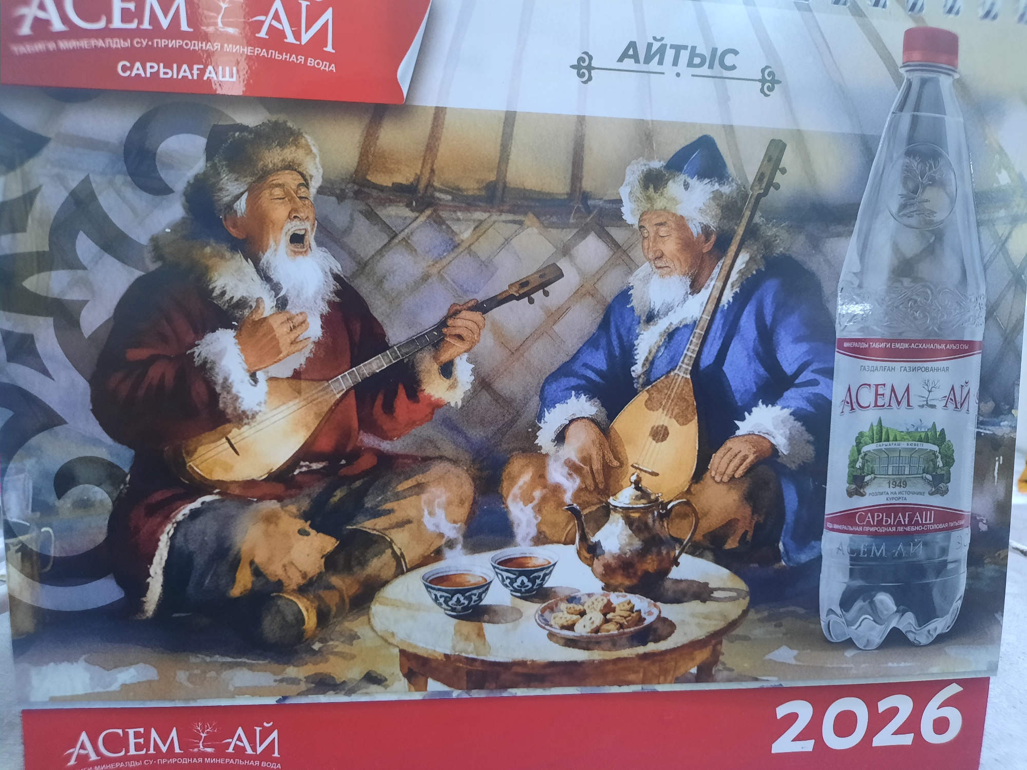 The image is a close-up shot of a calendar page depicting a watercolor painting of two elderly men in traditional attire playing musical instruments. One man, on the left, wears a dark red robe and a fur hat and is playing a stringed instrument. He has a white beard and appears to be singing with his eyes closed and mouth open. The other man, on the right, wears a blue robe and a fur hat and is playing a similar stringed instrument. He has a white beard and appears to be looking down.
In front of them is a round wooden table with two bowls of steaming tea and a small plate of cookies. A silver teapot sits on the table. The background shows the interior of a yurt, with woven mats and wooden poles visible.
To the right of the painting is a clear plastic bottle of "ACEM AI" mineral water, with labels in Cyrillic script. The bottle has a red cap and a label that shows a building with trees and the text "1949 РОЗЛИТА НА ИСТОЧНИКЕ КУРОРТА". Below this, the label states "САРЫАГАШ".
At the top of the calendar page, there are red and white text elements that say "ACEM AI" and "САРЫАГАШ". The text "АЙТЫС" is also visible on the top right.
At the bottom of the calendar page, prominently displayed in large white numerals, is the year "2026".
The text on the bottle and the calendar page indicates that the mineral water is from the Saryagash resort. While the image itself does not explicitly show the city of Zhanaozen, Kazakhstan, the brand "ACEM AI" and the resort "Saryagash" are associated with Kazakhstan. Saryagash is a resort town in Kazakhstan known for its mineral water springs, located in the Turkestan Region, which is geographically distinct from Zhanaozen, a city in the Mangystau Region. Therefore, while the image promotes a product and location within Kazakhstan, it does not directly depict Zhanaozen.
