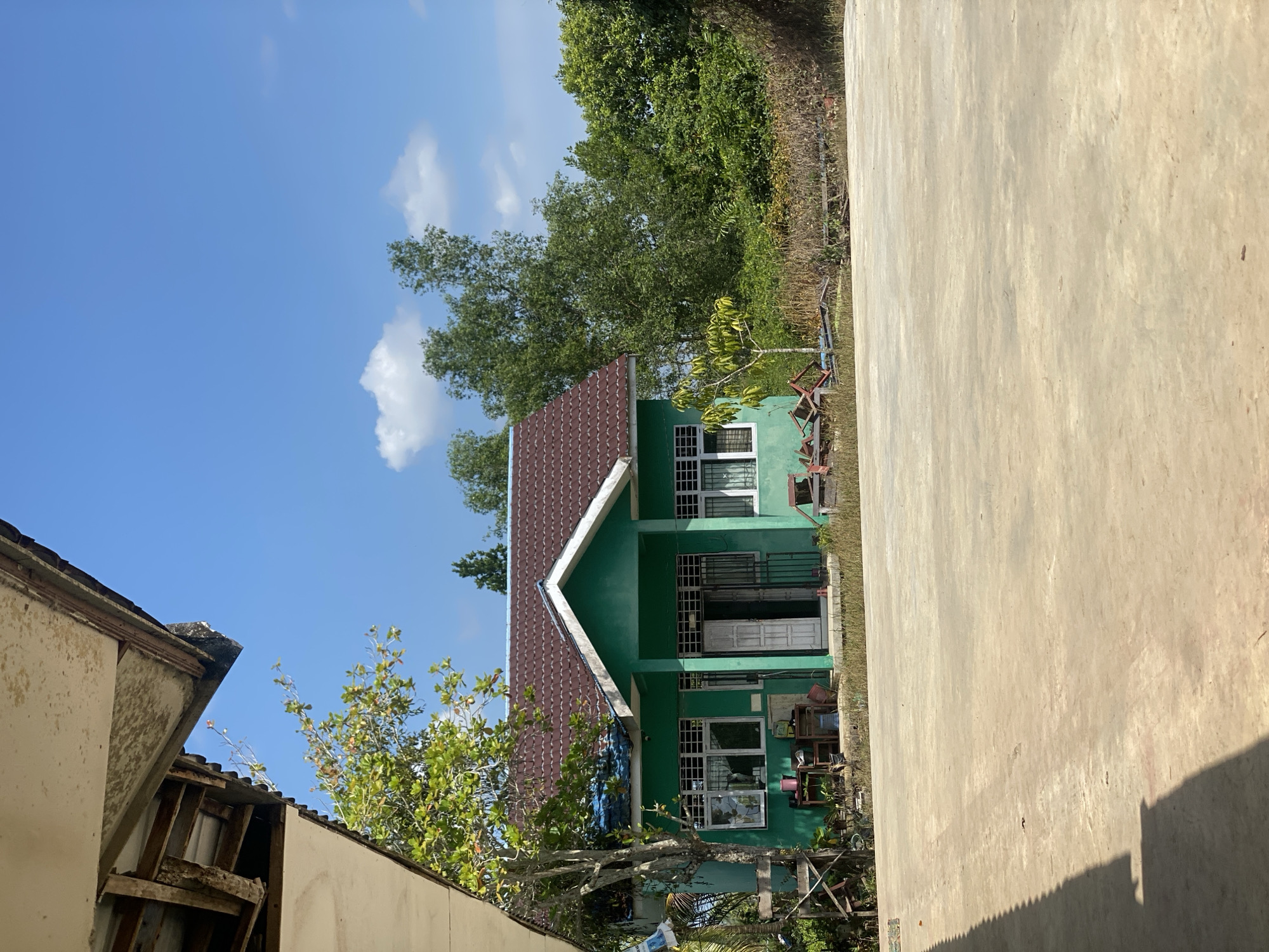 A bright, sunny day is depicted in Handil Baru, Indonesia. The image features a small, green building with a brown, tiled roof. The building has several windows with white frames and grates, and a double door entrance. In front of the building is a large, paved area. To the left, a portion of another building's roof and ceiling is visible, showing some signs of wear. The background consists of lush green trees and foliage under a clear blue sky with a few scattered white clouds. Some debris and plants are scattered around the base of the green building and the edge of the paved area. The scene appears to be an outdoor space, possibly a school or community building, in a tropical environment.