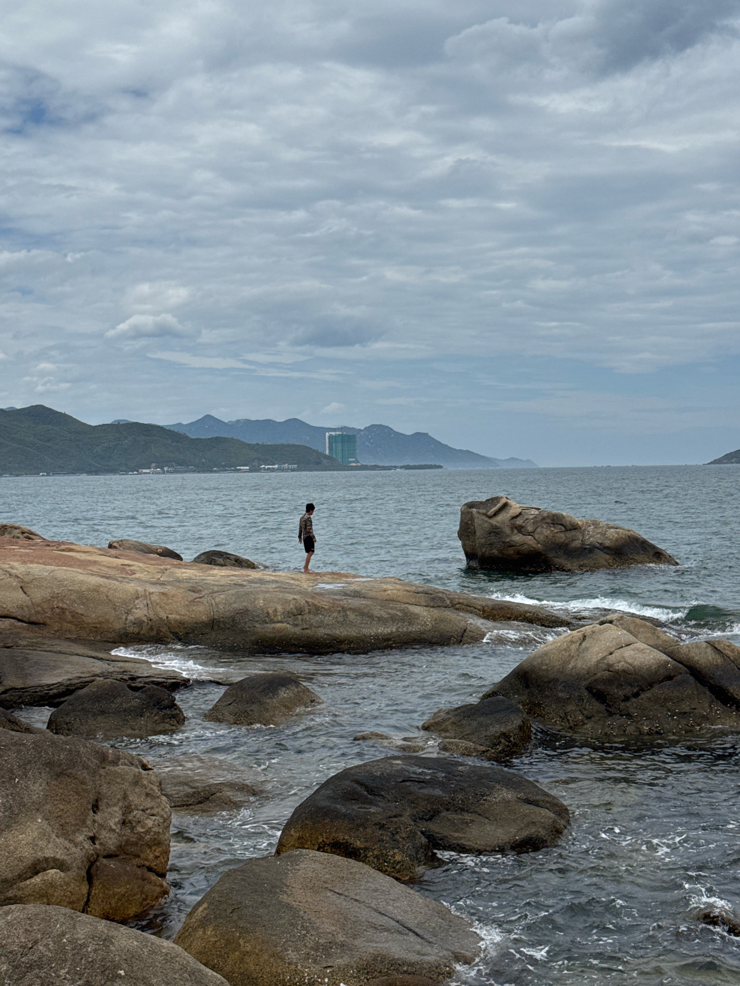 A lone individual stands on a rocky outcrop overlooking the sea, facing away from the camera. The person is wearing a patterned shirt and dark shorts. The foreground is dominated by large, weathered rocks, some partially submerged in the gently lapping waves. In the mid-ground, the sea extends to a distant coastline marked by green, rolling hills and mountains. A large, modern building with a green facade is visible nestled among the hills on the right side of the distant landscape. The sky is overcast with diffused light. The scene is likely a coastal area in or near Nha Trang, Vietnam, possibly a scenic spot known for its rocky shores and views of the city's outskirts.