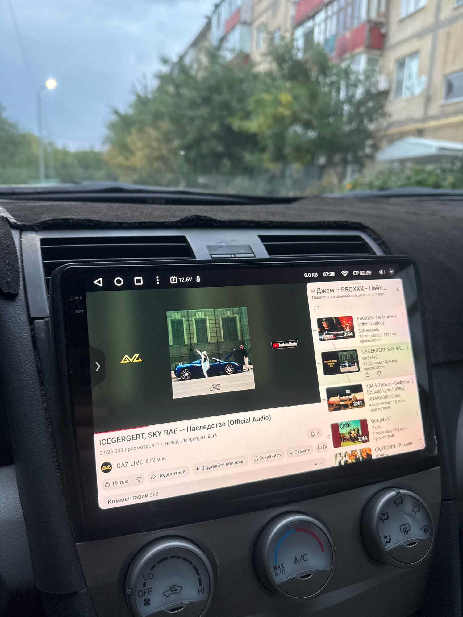 The image shows the interior of a car in the city of Aktobe, Kazakhstan. A large touchscreen display is mounted on the dashboard, showing a YouTube music interface. The screen displays a song titled "ICEGERGERT, SKY RAE – Наследство (Official Audio)" with accompanying metadata such as view count and upload date. Next to the main video are suggested videos, one of which is also titled "PROXXХХ – Найтвижн (official video)". The car's climate control knobs are visible below the screen. Through the car's windshield, a street scene is visible, with buildings and trees, suggesting an urban environment. The weather appears overcast.