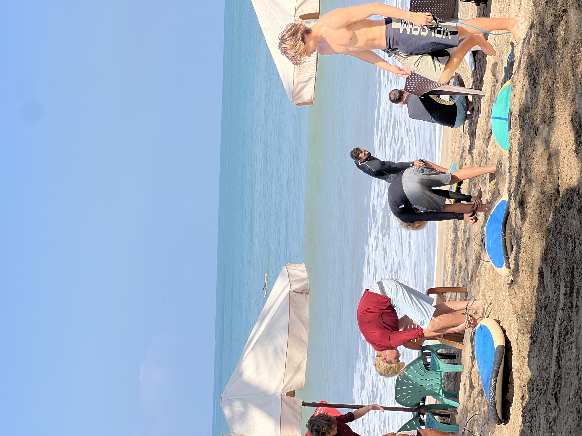 In Kuta, Indonesia, on a sunny day, a group of surfers are gathered on the sandy beach. The ocean, a vibrant turquoise, stretches out to the horizon under a clear blue sky. In the foreground, several surfboards are laid out on the sand, some blue and white, one green and yellow. One surfer, a young man with blonde hair, is bending down, attaching a leash to his surfboard. He is wearing a red long-sleeved rashguard and light-colored shorts. Another surfer, wearing a black wetsuit, is also bent over, appearing to adjust his gear. A shirtless young man with blonde hair stands on the right, looking towards the water, wearing dark shorts with a logo. In the background, two people are seated on chairs under a large, cream-colored umbrella. Further out in the ocean, a lone figure is lying on a surfboard. The scene captures the relaxed atmosphere of a surf lesson or a day of surfing at the popular Kuta beach.