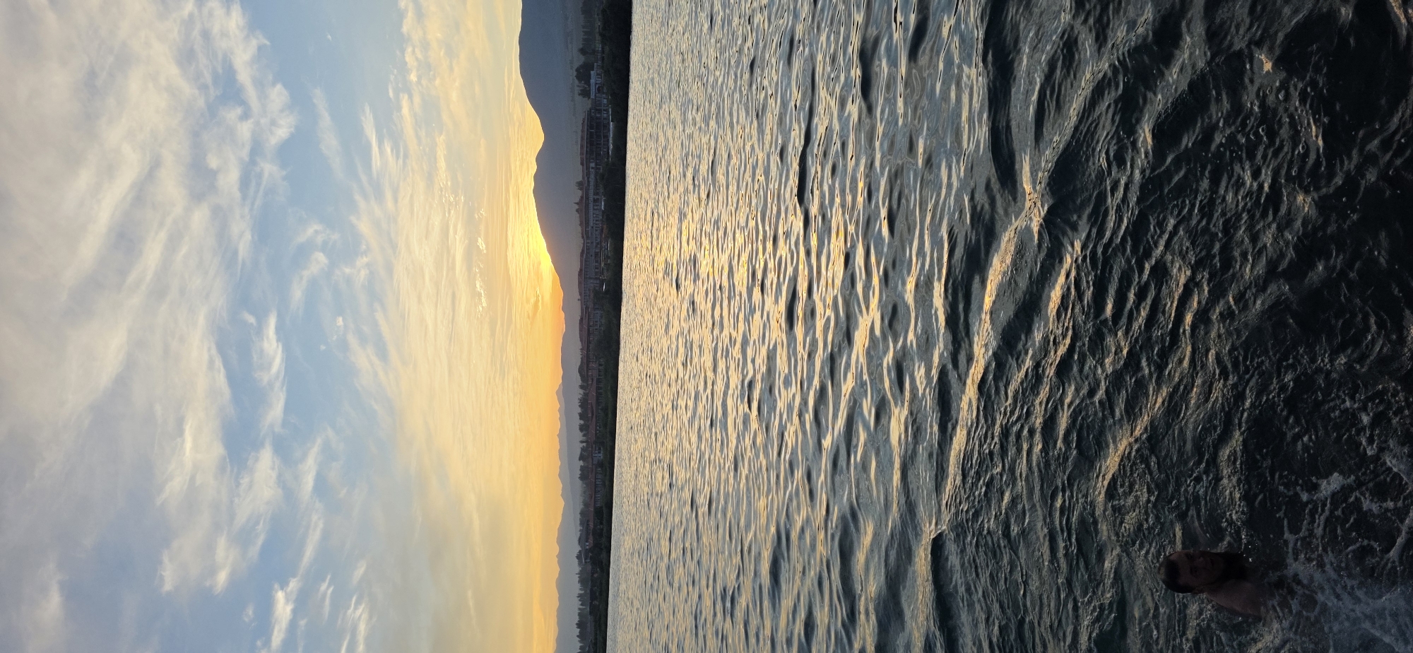 A man is swimming in dark water, with waves and reflections of the sunset visible. In the background, a city with buildings and mountains can be seen against a sunset sky with clouds. The location is identified as Choktal, Kyrgyzstan.