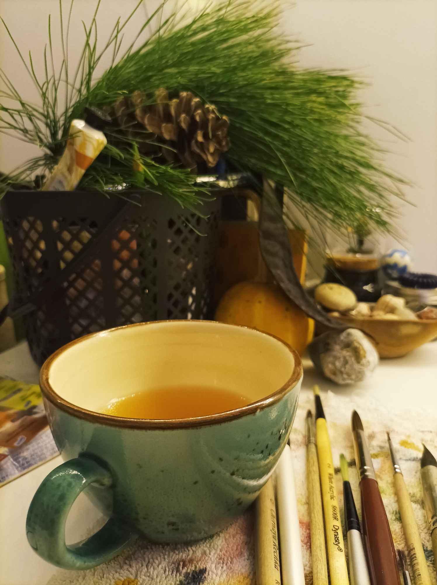 This image depicts an indoor still life scene, located in the city of Batumi, Georgia. The foreground features a teal-colored ceramic mug with an off-white interior and a brown rim, filled with an amber-colored liquid. The mug rests on a white, textured fabric, possibly a towel, exhibiting faint colored patterns. To the right of the mug, a collection of approximately ten artist's paintbrushes with varying handle colors (yellow, white, brown, natural wood) and brush types are laid horizontally on the same fabric. Some brush handles display partial text, including "White Acrylic" and "Dynasty."
In the midground, a black, latticed metal container holds evergreen branches, identified as pine or spruce, adorned with at least one visible brown pinecone. A small, light-colored, foil-wrapped object is partially obscured among the branches within the container. To the right of the container, a small, orange-yellow object resembling a mini pumpkin or gourd is present. Further right, a small wooden bowl contains multiple light-colored, smooth objects, consistent with pebbles or small stones. Additional indistinct decorative elements are partially visible in this area. A dark, curved object, possibly a metallic ornament or part of another container, is situated behind the gourd. The background consists of a plain, light-colored, out-of-focus wall or surface. No individuals are present in the scene, and no overt actions or interactions are depicted. The overall lighting is warm, suggesting an artificial light source or ambient indoor conditions.