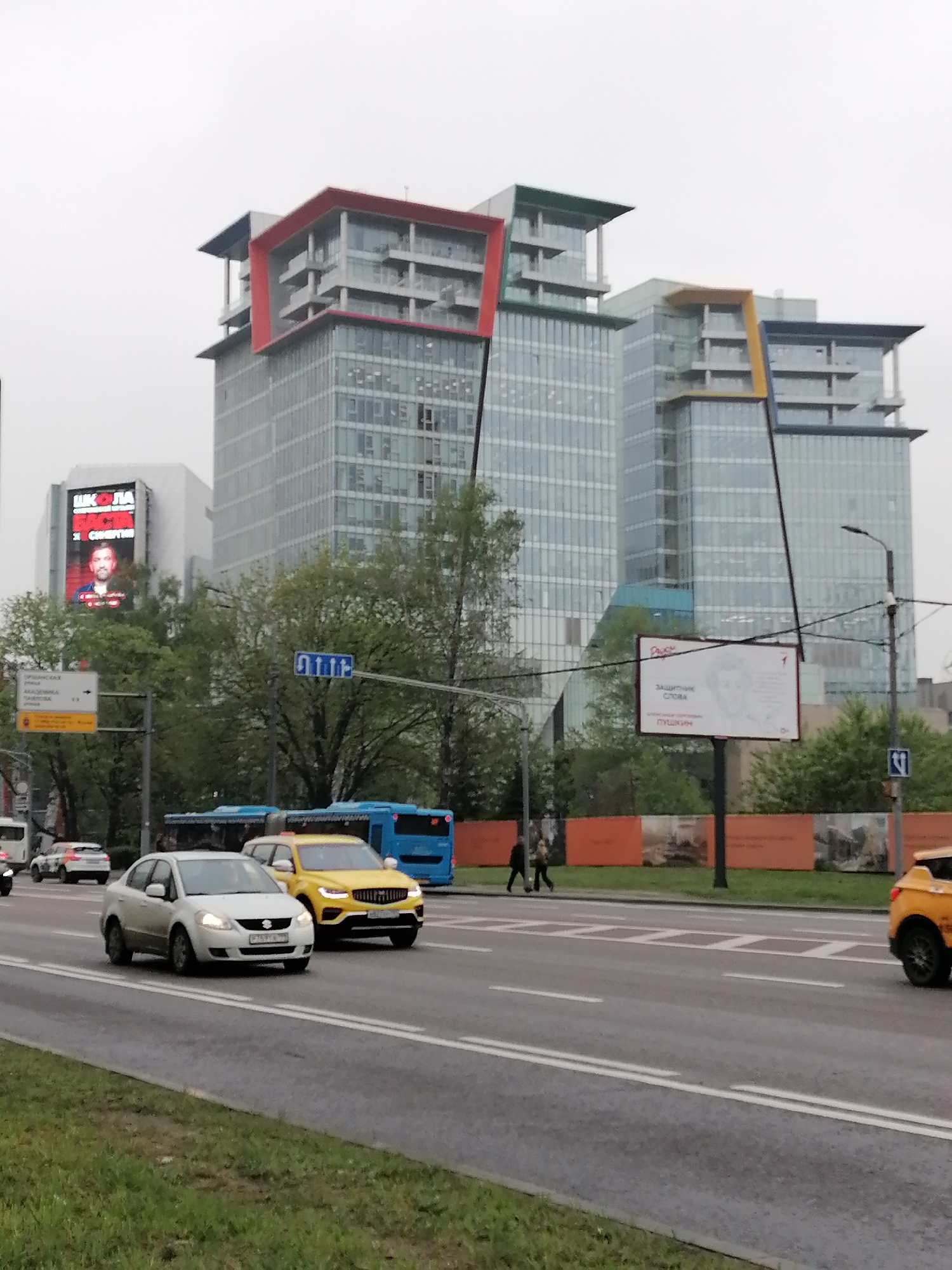 The image depicts a street scene in Moscow, Russia, under an overcast sky. Modern office buildings are visible in the background, characterized by glass facades and colorful architectural details on the upper levels (red, green, yellow). A digital billboard displays an advertisement with a man's face.
In the foreground, a multi-lane road carries vehicular traffic, including a white Suzuki SX4 and a yellow crossover SUV. Behind them are several blue transit buses. The median strip separating the lanes is green, with trees and signs. A billboard advertising "Defender of the Word Pushkin" is also visible on the median. Pedestrians are walking along the sidewalk. The street has white painted lines.