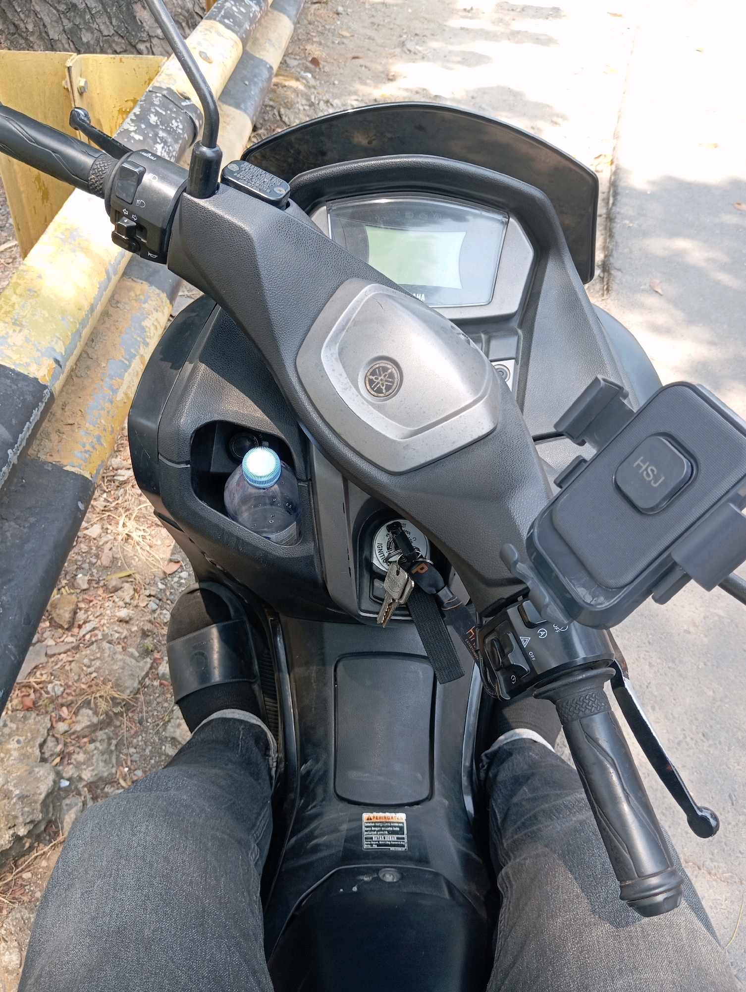 The image shows a first-person perspective of someone sitting on a black motorcycle, likely in Semarang, Indonesia. The motorcycle's handlebars, instrument panel, and the rider's legs are visible. A phone mount with "HSJ" printed on it is attached to the handlebars. A bottle of water is in a compartment to the left of the instrument panel. The rider is wearing dark grey jeans and black sandals over black socks. The background consists of a yellow and black striped barrier, rough ground with stones and dry leaves, and asphalt. Sunlight casts shadows, indicating it is daytime. A warning sticker is visible on the motorcycle's bodywork.