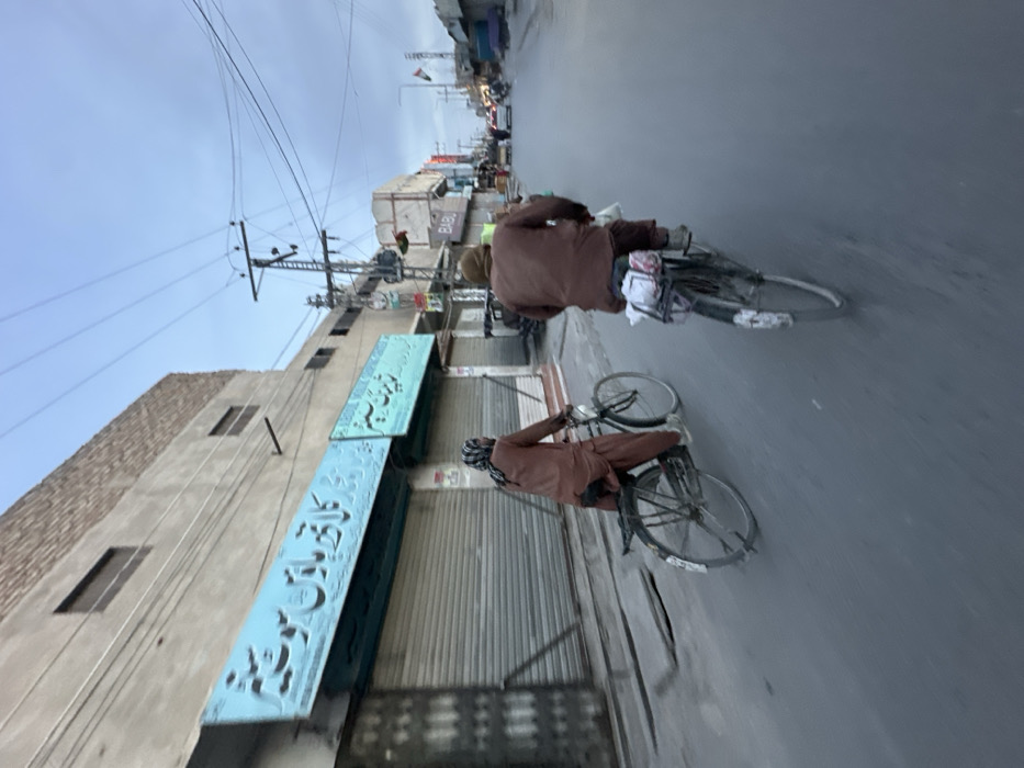 The image displays an urban street scene in Quetta City Tehsil, Pakistan, under an overcast sky. Two individuals, both appearing male and wearing brown traditional South Asian attire (shalwar kameez), are seen riding bicycles away from the camera. The individual in the foreground is on an older model bicycle with items secured to its rear rack. The individual to their left, slightly further back, is also on an older model bicycle and wears a patterned head covering.
On the left side of the street, a two-story building features a light blue signboard with Urdu script, which reads "کالا برتن ہاوس سوفتیر" (Kala Bartan House Software), positioned above multiple closed metal roll-down shop shutters. The upper part of this building is constructed with concrete and exposed brick. Numerous utility poles with a dense network of overhead electrical wires are visible along the street. Further down the road on the right, a building displays a sign with the name "BABU". In the far distance on the right, a flag with distinct black, red, and green horizontal stripes is flown from a pole, and sparse vehicular traffic is present on the road. The street is a paved asphalt surface.