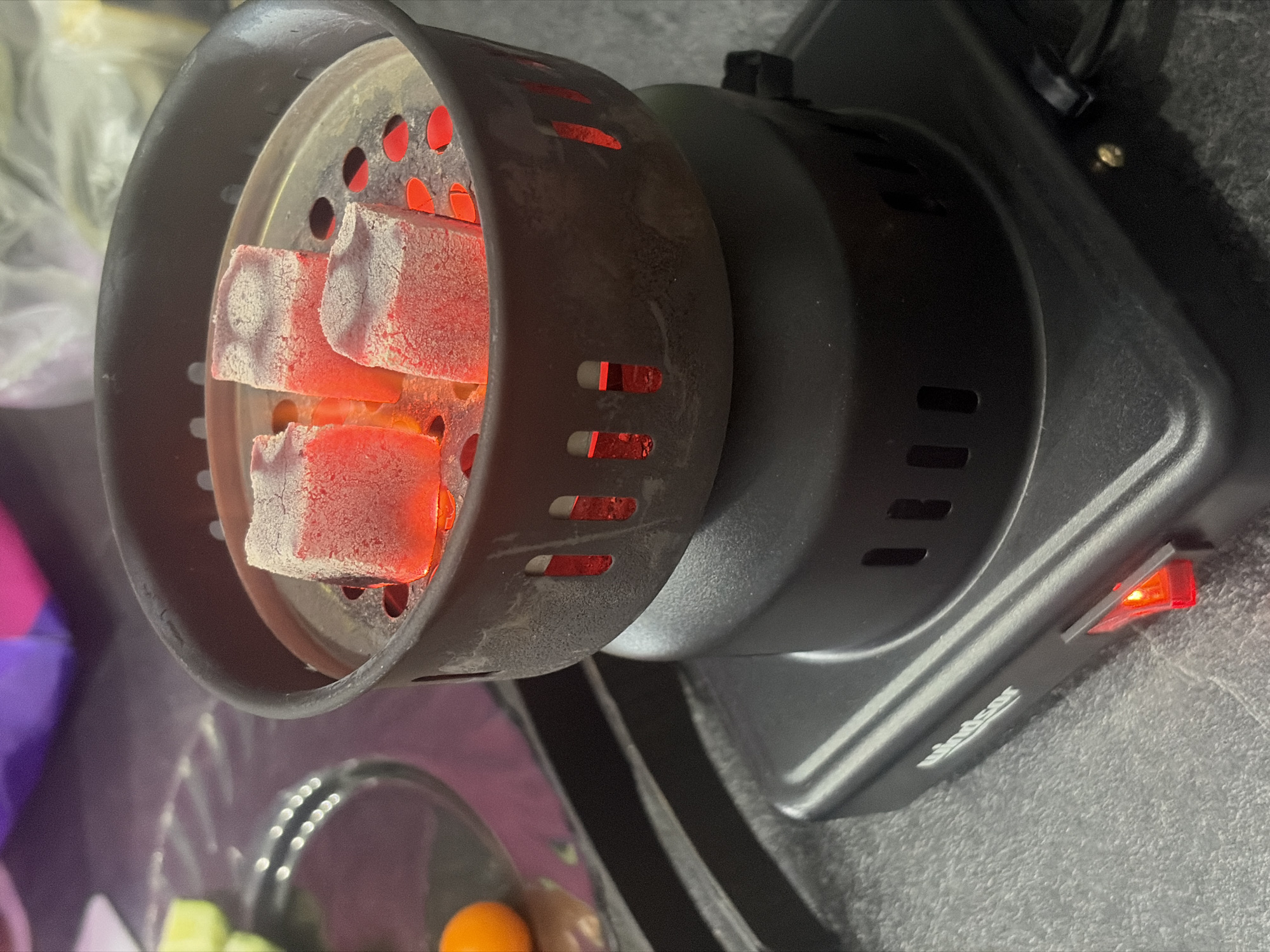 The image displays a close-up, slightly angled view of a black electric charcoal burner, actively heating several rectangular charcoal cubes. The charcoal pieces are glowing an intense red, signifying they are fully ignited and prepared for use, likely for a shisha or hookah. A thin layer of white ash or residue covers parts of the glowing charcoal. The metallic housing of the burner, which contains the coals, features various circular and elongated openings through which the bright red light from the charcoal is visible.
The main body of the charcoal burner is dark, and on its right side, a red rocker switch is illuminated, indicating the device is operational. The brand name "Windsor" is faintly visible in white script below the switch. The burner is placed on a dark, textured surface, possibly a countertop. The blurred background shows reflections and indistinct shapes, including what appears to be a clear plate holding pieces of fruit, such as an orange segment and some green items, suggesting an indoor environment. The lighting conditions imply an evening or nighttime setting, highlighting the glow of the charcoal and the illuminated switch. No individuals are present in the image.