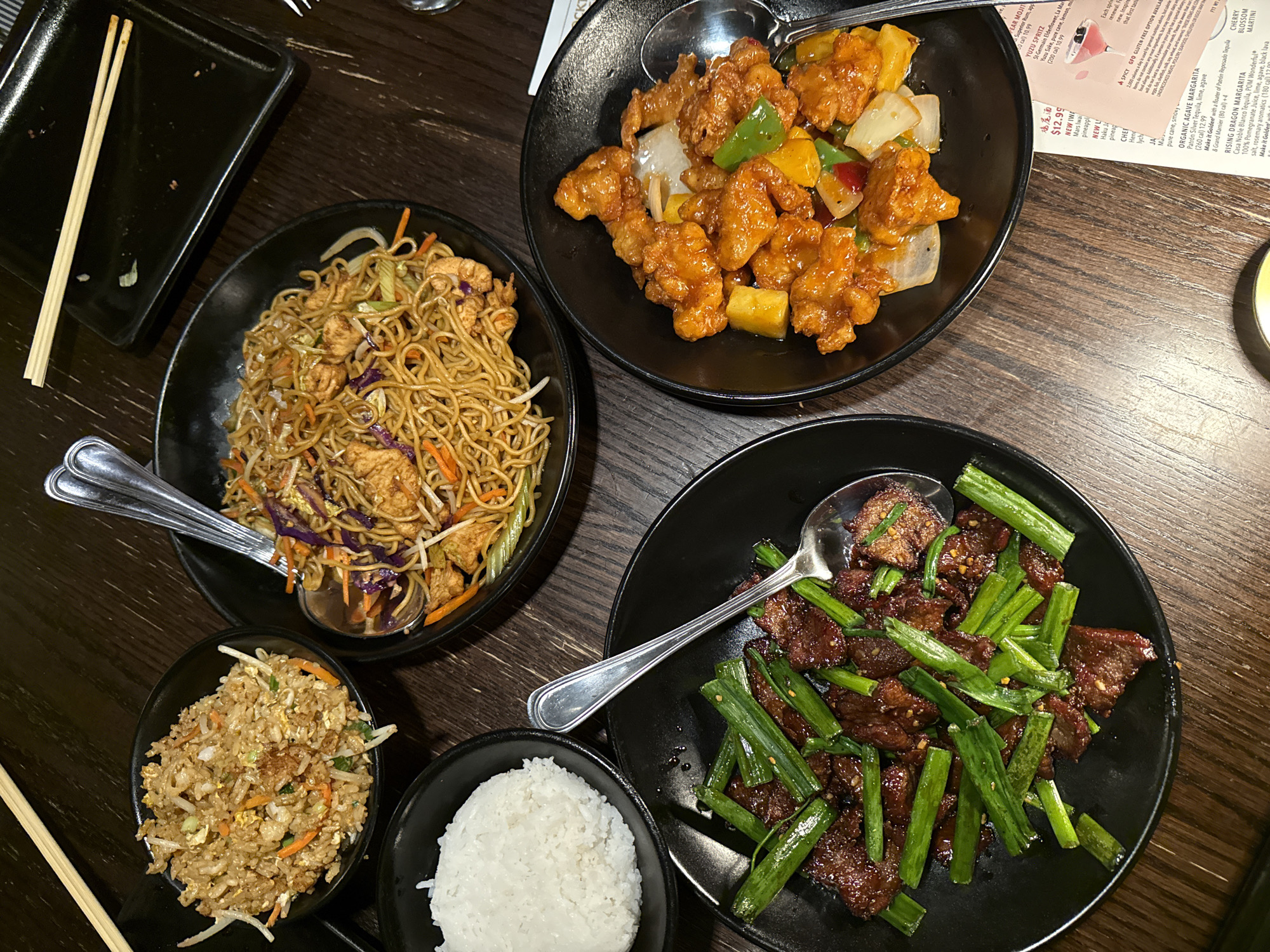 The image shows a table setting in a restaurant, featuring an assortment of Asian-style dishes. The dishes include chow mein, sweet and sour chicken, stir-fried beef with scallions, fried rice, and a bowl of white rice. Silver serving spoons are placed in each dish, except the bowls of fried and white rice. There are chopsticks on a small black square plate. A menu is partially visible in the upper right corner. The scene is located in a restaurant in Chula Vista, California.
