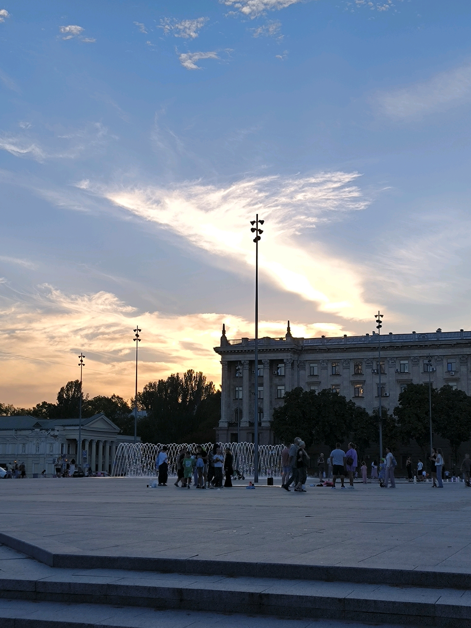 The image depicts an outdoor public square in Mykolaiv, Ukraine, likely taken during the late afternoon or early evening. The sky features a dramatic display of clouds, with streaks of white and orange suggesting the setting sun. Dominating the background is a large, classical-style building with columns and multiple windows. In the mid-ground, a fountain is active, with a cluster of people standing around it. Further left, another smaller classical building is visible. Several tall lamp posts with multiple lights are positioned throughout the square. In the foreground, there is a wide, paved area with stone steps leading up from the lower edge of the frame. A group of people are scattered across the square, some appearing to be walking or standing in small groups. The overall atmosphere suggests a public gathering space being used for leisure.