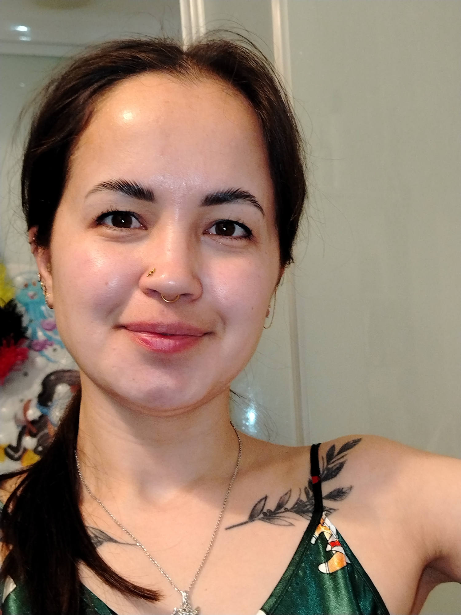 A close-up selfie photograph features a woman with dark brown hair pulled back into a low ponytail. She has light skin, brown eyes, and defined eyebrows. She is wearing a nose ring and small earrings. The woman is smiling softly.
She has a leafy branch tattoo on her left shoulder. A silver necklace with a pendant hangs around her neck. She is wearing a green top with an indiscernible pattern. The background is a neutral-toned wall.