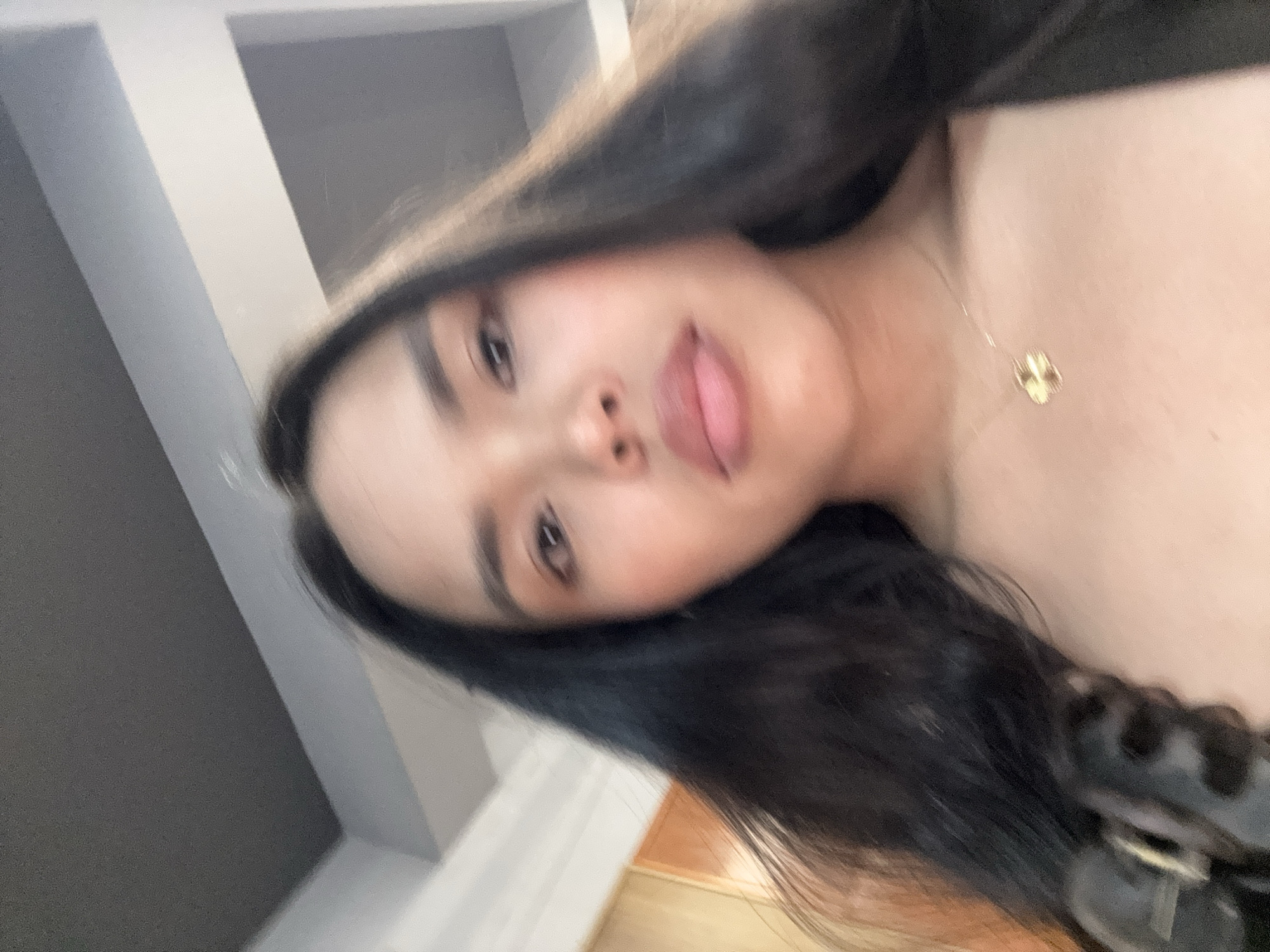 The image is a close-up, blurry selfie of a woman with dark hair. She is wearing a necklace with a four-leaf clover pendant. The background is out of focus, showing parts of a ceiling and a wall. The location of this image is not determinable from the provided visual information.