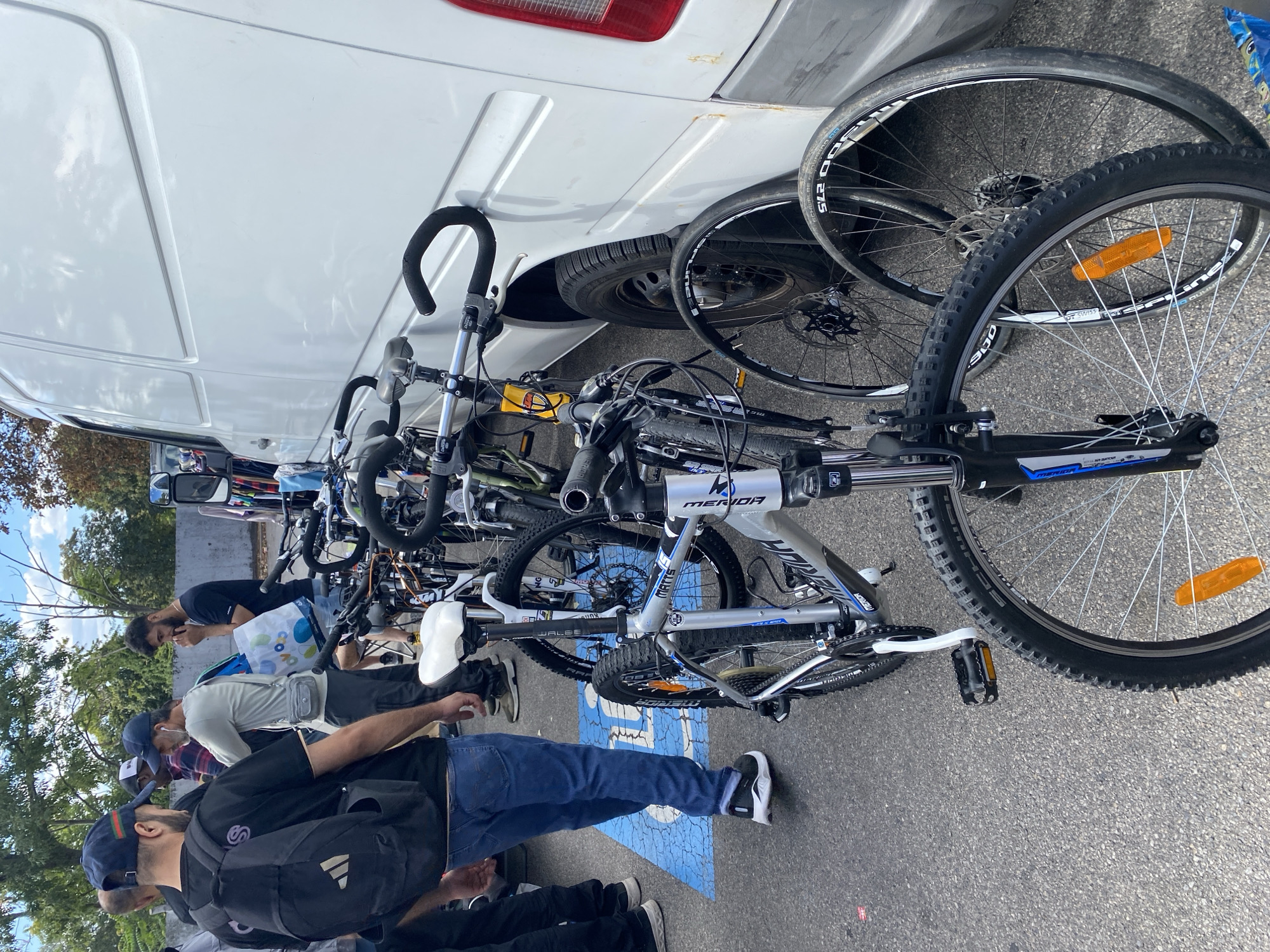 The image depicts an outdoor scene in Vienna, Austria, near a white van, with several bicycles and a group of men. In the foreground, a silver and blue mountain bike is prominently displayed, with its front wheel and handlebars visible. To its left, another mountain bike with a white seat and blue accents is partially visible. Further back, additional bicycles, some with yellow frames, are lined up. A white van, the side of which is visible, is parked next to the bicycles. Several men are present in the scene, some appearing to be looking at or interacting with the bicycles. One man in the foreground wears a black t-shirt, blue jeans, and a black backpack. Another man, facing away from the camera, wears a grey t-shirt and dark pants. A third man in a black t-shirt and shorts stands further back. The ground appears to be asphalt, with a blue and white disabled parking symbol visible under one of the men's feet. The background shows trees and a cloudy sky, suggesting an outdoor market or a similar event.
