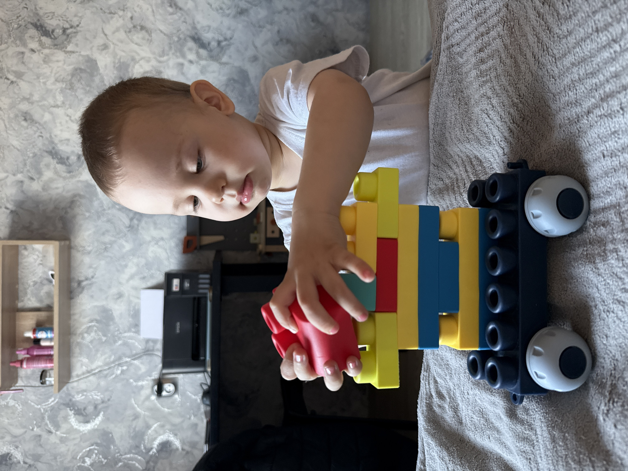 A young child wearing a white shirt is playing with large plastic building blocks on a textured surface. The blocks are various colors including red, green, yellow and blue. They are arranged to resemble a vehicle with navy blue wheels. The child is actively manipulating the blocks with their hands. In the background, there is a shelf with small containers, a printer, and a saw hanging on the wall. The location is Rusanovo, Russia, based on the available data.