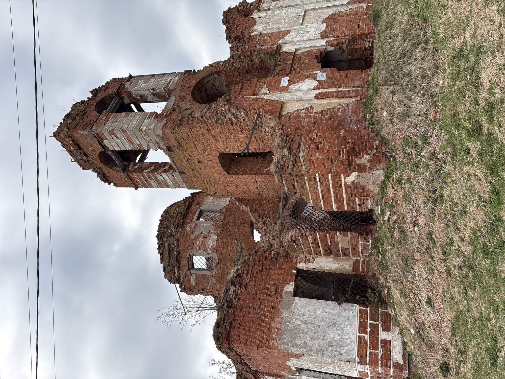 The image shows the ruined Church of the Intercession of the Holy Virgin, located in Staroe Rakomo, Russia. The church, constructed of red brick, is heavily damaged, with large sections of walls and roof missing. Patches of remaining white plaster are visible on the brickwork. Some window openings have remnants of metal grating. Vegetation, including grass and small trees, grows on and around the ruins. A utility line is visible against a cloudy sky.
