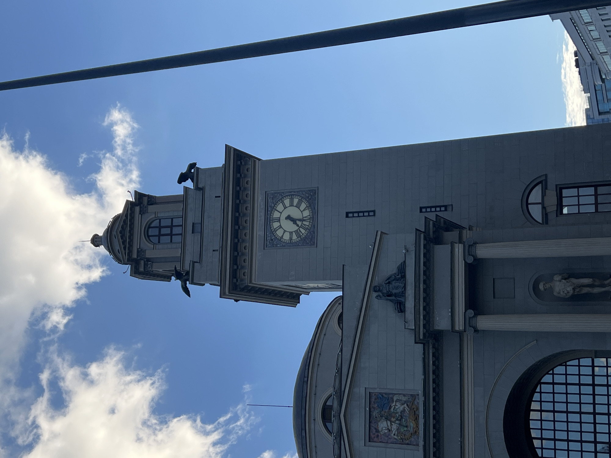 This media file captures a view of a grand building with a prominent clock tower against a bright blue sky with scattered clouds. The building appears to be made of stone or concrete and features classical architectural elements such as columns and ornate detailing. A large clock face is visible on the tower, indicating the time. Statues and decorative panels are integrated into the building's facade, adding to its imposing and artistic character. The perspective is looking upwards from a low angle, emphasizing the height and grandeur of the structure. A dark horizontal bar runs across the top of the frame, obscuring a portion of the sky and suggesting an overhead structure or obstruction. The overall impression is one of a significant civic or historical building, possibly a town hall or museum, in an urban setting. The bright sky suggests daytime, likely sunny weather. The image is oriented vertically, with the top of the building appearing on the left side of the frame. There is no visible text within the frame.