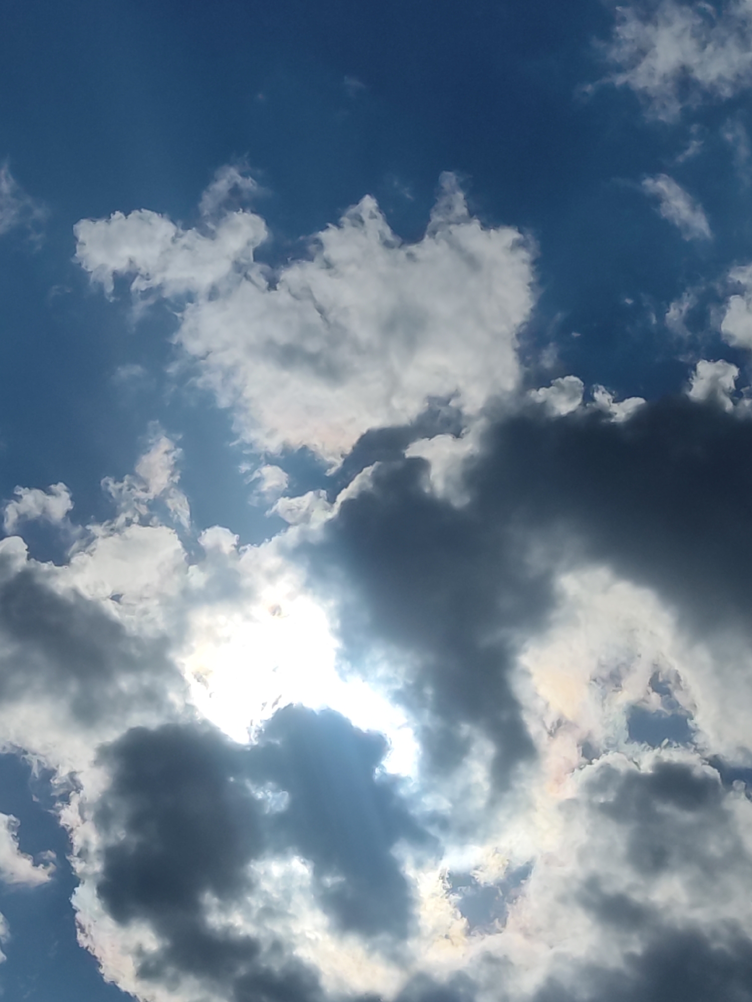 The image shows a partly cloudy sky in Chaikovsky, Russia. The sky is blue, with a mix of white, gray, and dark gray clouds. The sun is visible through a break in the clouds, creating a bright spot in the lower center of the image. There are no people or other objects visible in the image, apart from the clouds and sky. The environment appears to be a typical daytime scene.