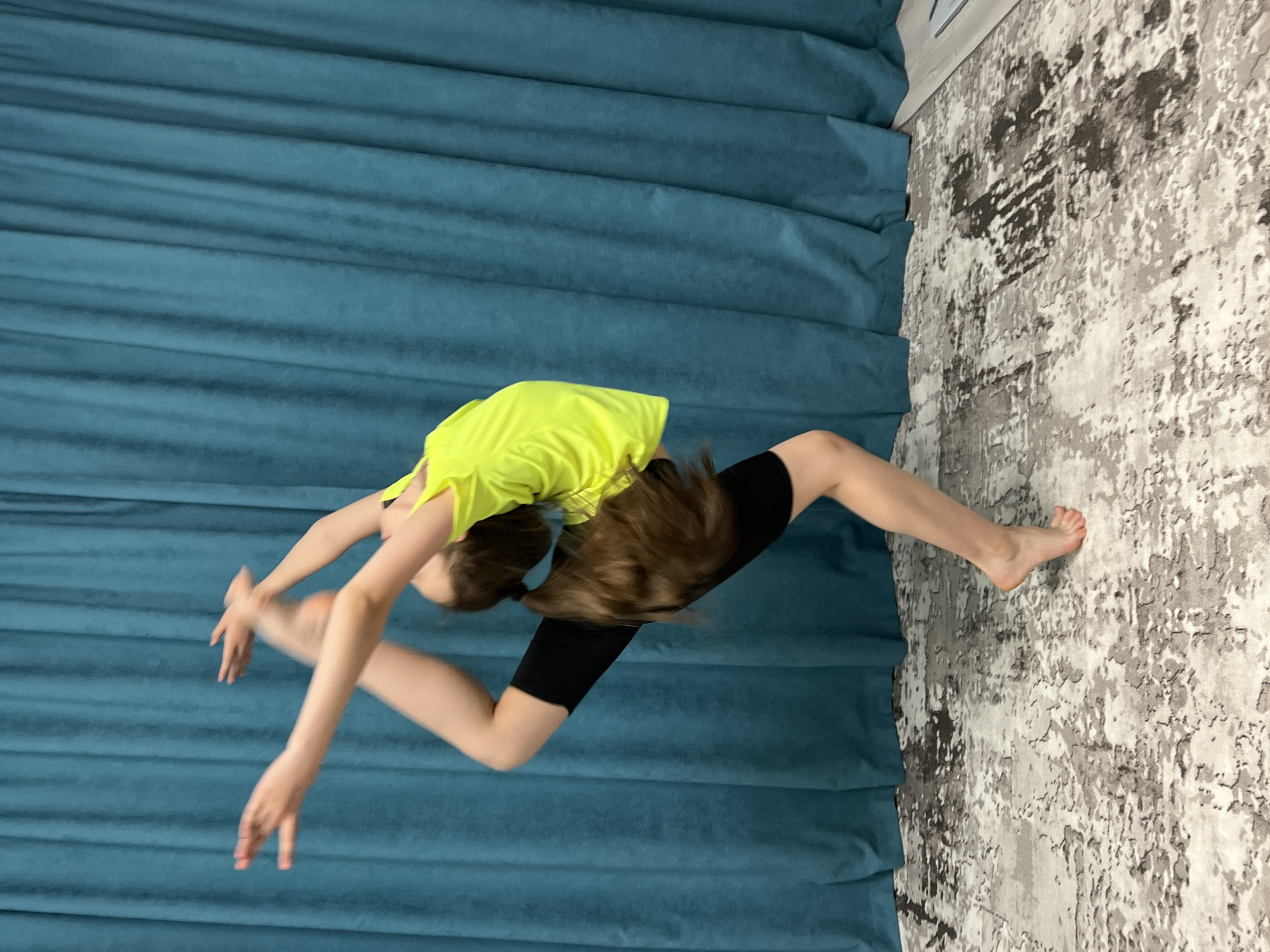 A person wearing a yellow top and black shorts is performing a backbend split in a room in Chaikovsky, Russia. The person's hair is brown, and their feet are bare. The background consists of a blue curtain on one side and a patterned wall or carpet on the other.