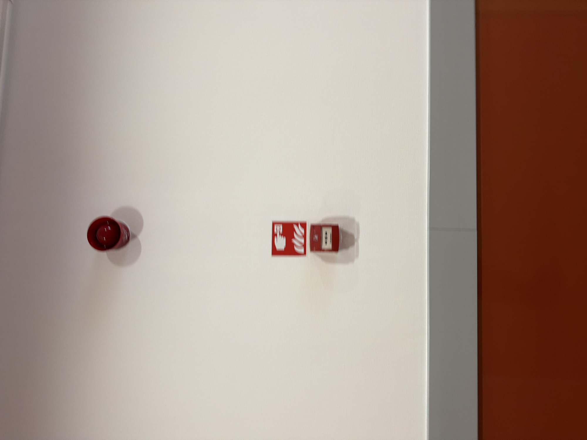 An indoor image shows a white wall with a gray trim running horizontally near the bottom and an orange surface below the trim. Mounted on the white wall are two red fire safety devices. At the top, slightly off-center to the right, is a red fire alarm siren. Below and to the left of the siren is a red fire alarm pull station with white graphics depicting flames and a hand. The pull station is mounted above a horizontal line that appears to be part of the wall's construction or decoration. No people are visible in the image. The scene is devoid of any context that would indicate its specific location within the city of Sofia, Bulgaria.