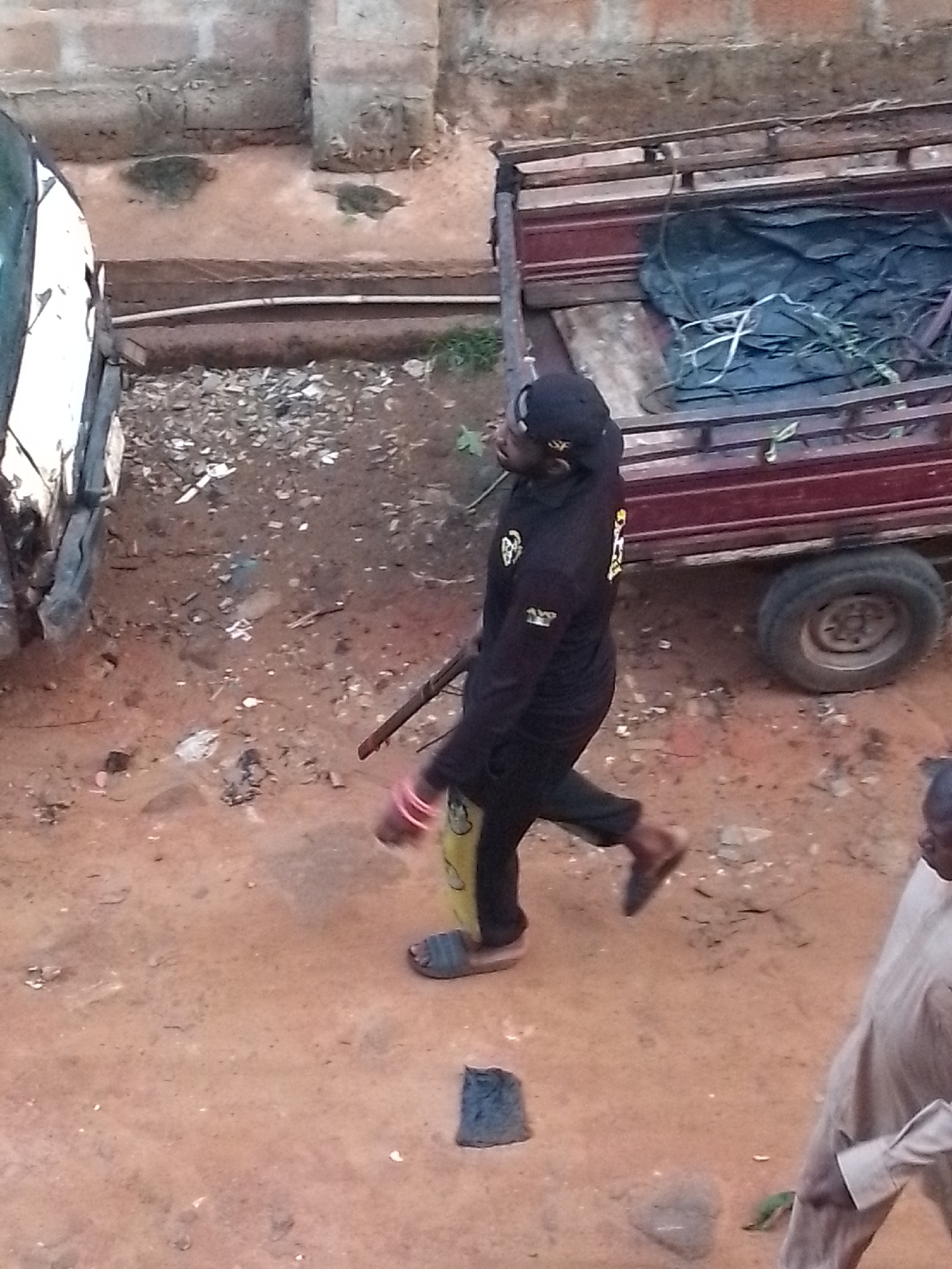 The image shows a man in dark clothing and a baseball cap walking in a dirt yard. He is carrying a long stick and wearing sandals. Next to him is a trailer with a black tarp inside. To the left, part of a white vehicle is visible. The location appears to be a yard or unpaved area possibly in the city of Unknown, Unknown. Another man is partially visible on the right.