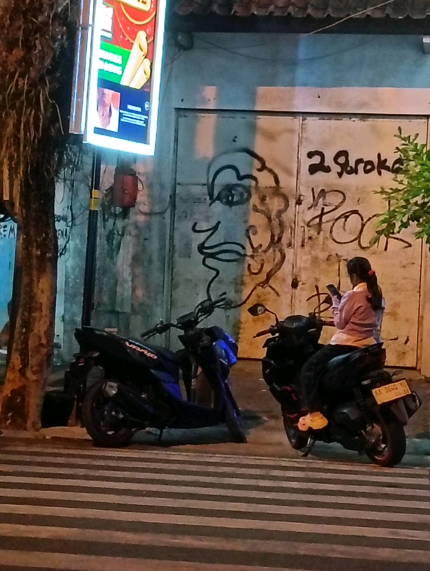 In the city of Purworejo, Indonesia, a scene unfolds on a street at night. A bright, illuminated sign displaying an advertisement for what appears to be food, possibly crepes or pastries, hangs on the left side of the frame. Below the sign, a red electrical box is mounted on a dark pole. Adjacent to this, a tall, rough-textured tree trunk stands, its branches bare and wispy.
Dominating the background is a large, textured wall with two vertical panels, likely a garage door or similar structure. This wall is adorned with graffiti. On the left side of the wall, a large, stylized outline of a human face is sketched in black. To the right of the face, the words "2 Stroke" are spray-painted in dark brown, followed by what appears to be "POK" written in a similar style.
Parked on a striped crosswalk in the foreground are two scooters. The scooter on the left is dark blue and black. The scooter on the right is black and is occupied by a person. This individual, a woman with dark hair tied in a ponytail, is wearing a light pink long-sleeved shirt and dark pants. She is seated on the scooter and actively using her mobile phone, looking down at the screen. Her feet are clad in bright yellow shoes. The license plate of her scooter is visible, reading "AA 3640 YU". A small green plant with visible leaves is growing on the right side of the frame, near the wall. The overall lighting suggests nighttime, with artificial light sources illuminating the scene.