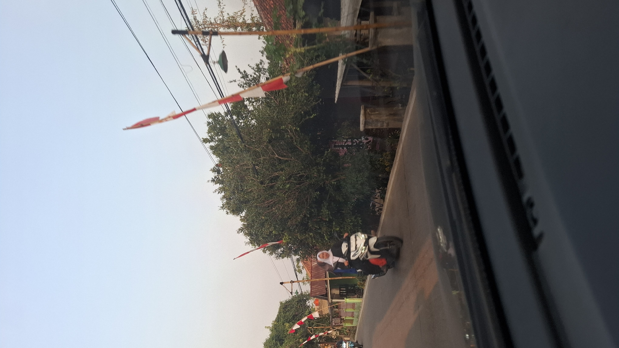 In Cibinong Hilir, Indonesia, a motorcycle carrying two individuals travels down a paved road. The motorcycle is white and the passengers are dressed in modest attire, one wearing a hijab. The scene is characterized by a clear sky and lush green trees lining the road. Several red and white flags, likely Indonesian flags, are visible, mounted on poles. Some of these flags appear to be tattered. Homes with corrugated metal roofs are situated behind the trees. A street lamp is also visible. The perspective suggests the photo was taken from a moving vehicle, with the dashboard visible in the foreground.