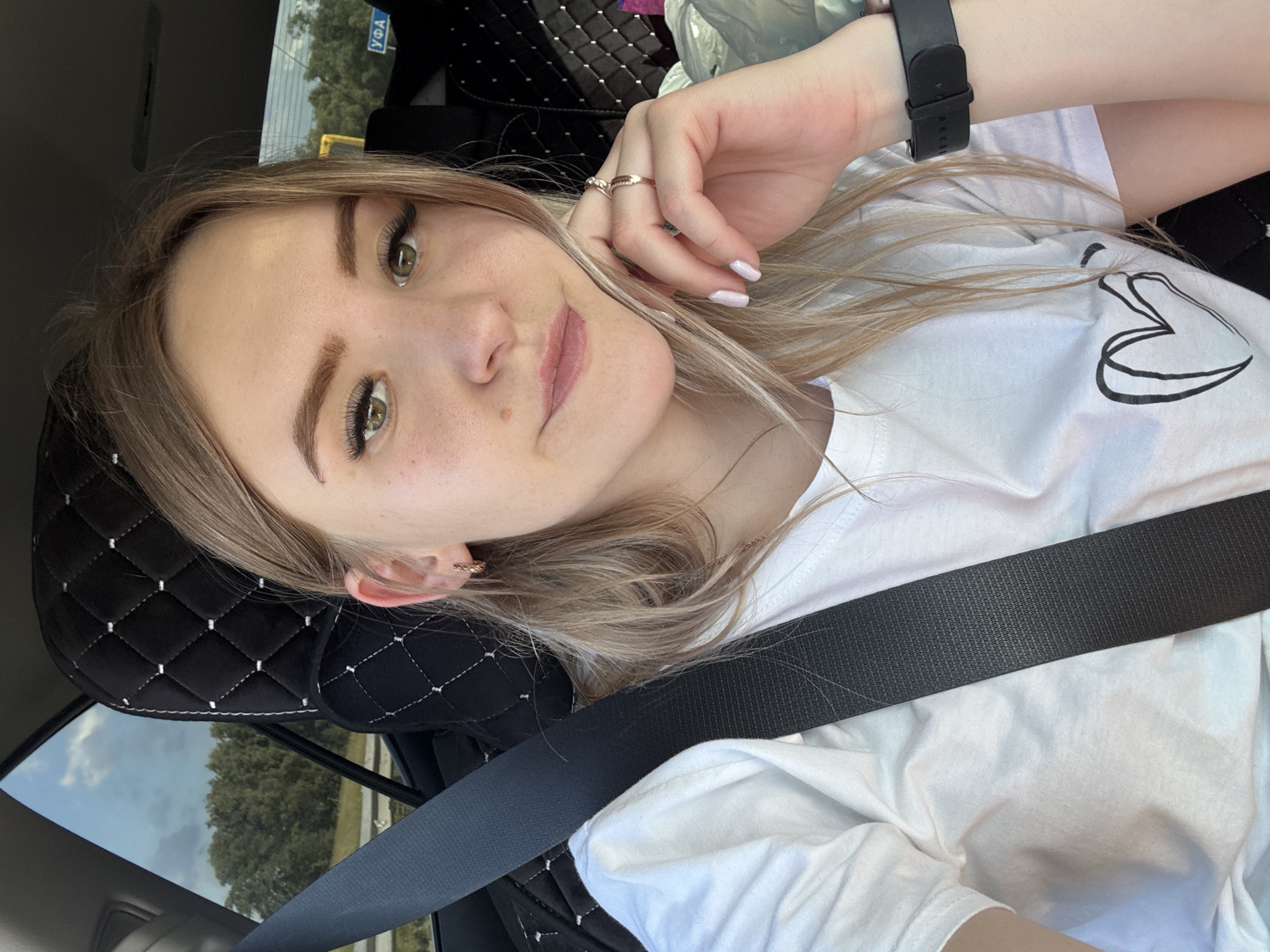 A young woman with blonde hair is shown in a selfie taken from a slightly high angle. She is wearing a white t-shirt with a black heart outline design on the right side of her chest. A black seatbelt is diagonally across her chest. Her left hand is resting on her cheek, and she is wearing a ring on her index finger and a watch on her left wrist. Her eyelashes are long and dark, and she has a light makeup application. Her lips are slightly parted, showing a pink lipstick color. A mole is visible on her cheek. The background suggests she is in a car, with a quilted black headrest visible behind her. Through a window on the left, trees and a cloudy sky are visible. A blue sign with white Cyrillic text and a yellow horizontal bar is also visible in the background. The overall impression is a casual, daytime selfie.