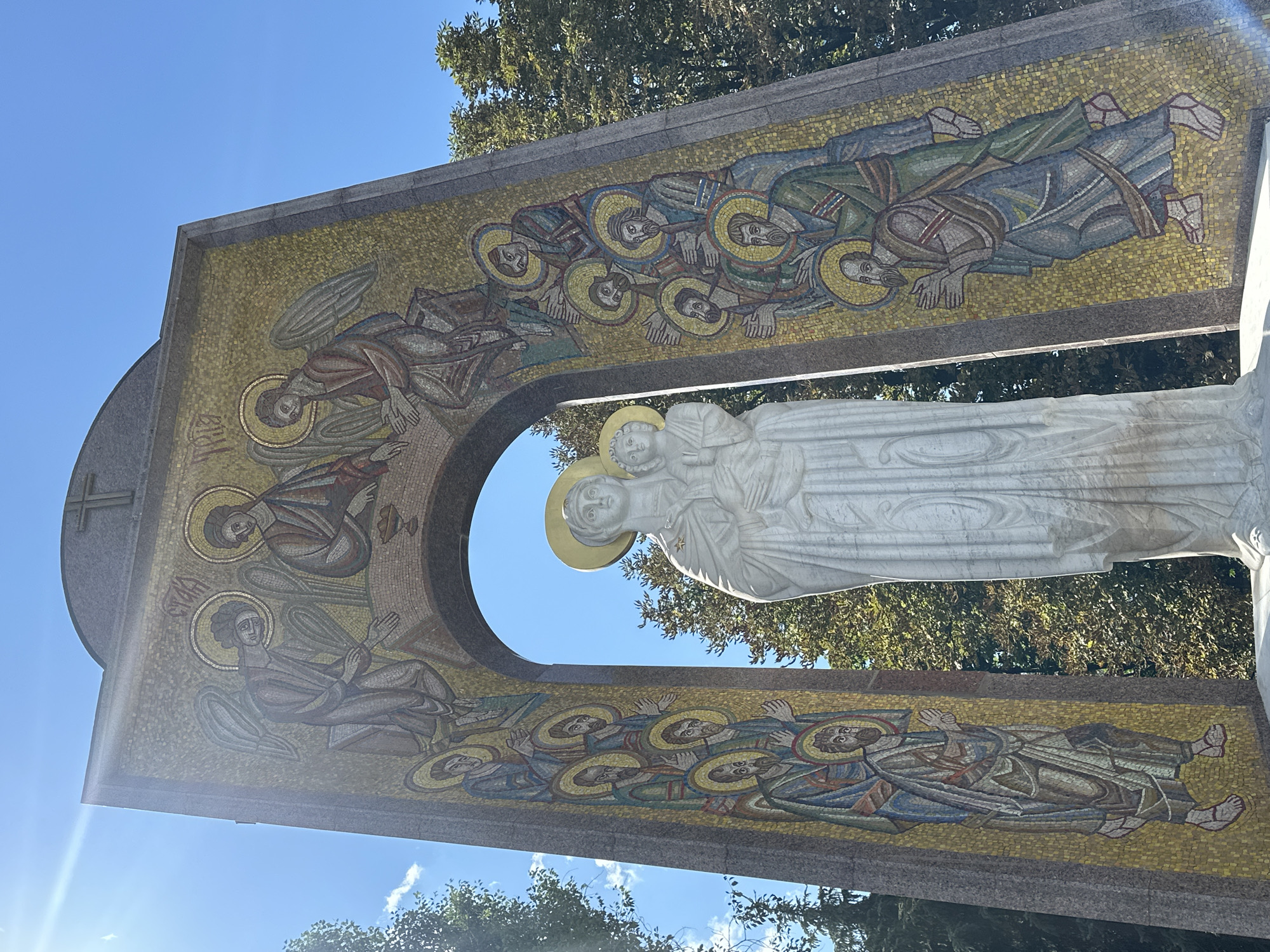 The image shows a large, arched monument featuring a mosaic on its side panels and a white marble statue of the Virgin Mary and Child at its center. The mosaic depicts religious figures, including angels and saints, arranged in rows and around the archway. The statue is positioned within the arch, with Mary holding the infant Jesus. The monument is set against a bright blue sky, with sunlight glinting off the mosaic and creating a lens flare in the upper left corner. Trees with green foliage are visible behind and to the sides of the monument. The monument is located in Raivka, Ukraine.