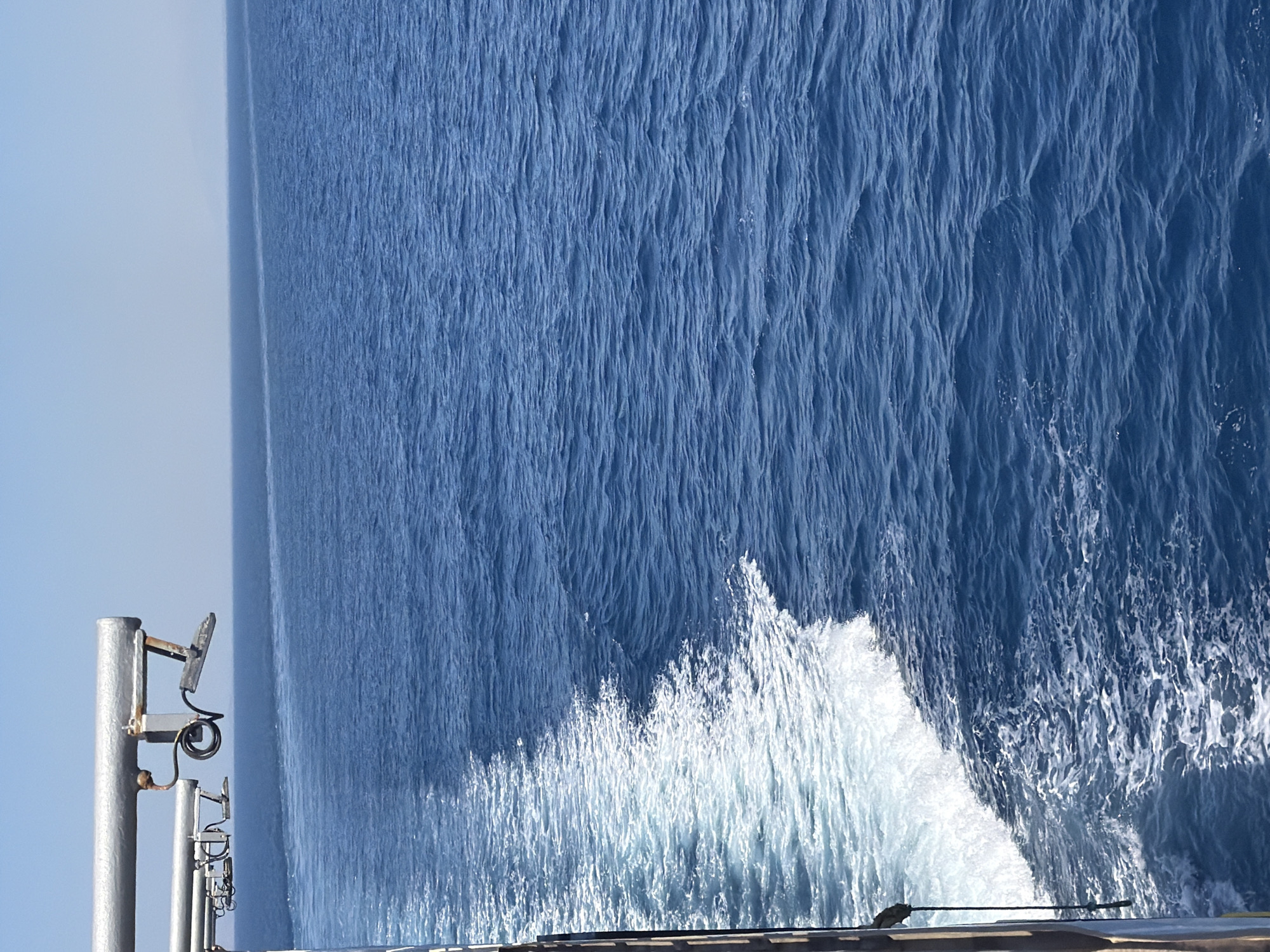 The image depicts a view from the side of a vessel moving through the sea, creating a wake. The water is a deep blue with white foam trailing behind. In the upper left corner, there are metallic structures extending from the vessel, including pipes and what appear to be sensors or equipment attached to them. The horizon is visible in the distance, with a clear, light blue sky above. The image captures the environment of open water, likely near the coast of the Municipal Unit of Kastro - Kyllini, Greece, given the context of a vessel at sea in that region. There are no people visible in the image.