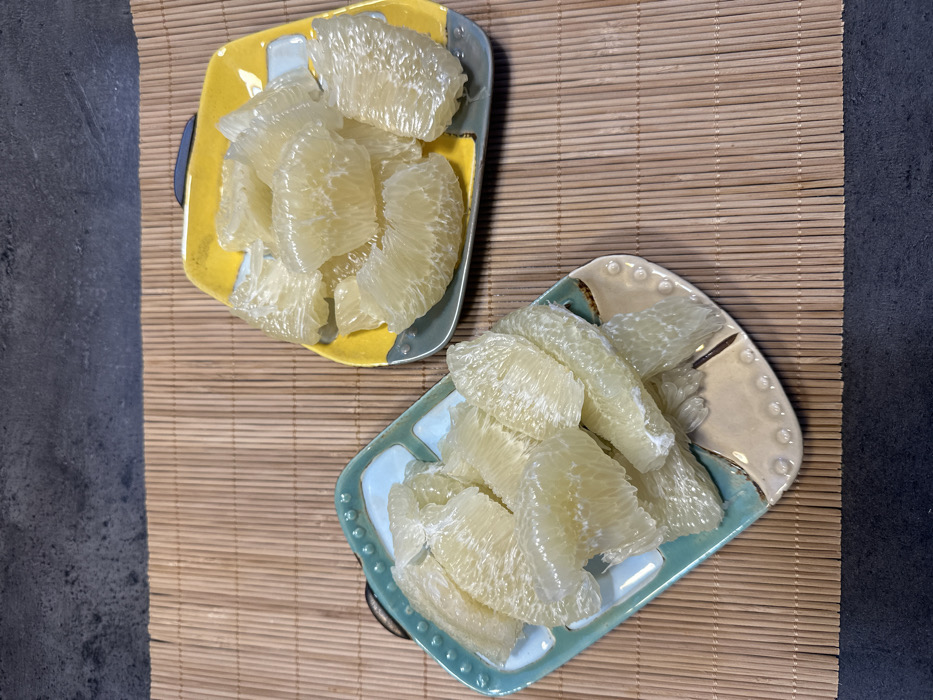 Two ceramic plates, each containing multiple segments of peeled pomelo, are centered on a light brown bamboo mat. The mat is positioned on a dark grey, textured surface. The plate on the left is teal and beige, while the plate on the right is yellow and grey; both are rectangular with rounded corners. The pomelo segments are pale yellow-green and appear moist. No individuals or specific actions are depicted. The scene is an indoor environment. The precise location in Tbilisi, Georgia, cannot be determined from the visual information.