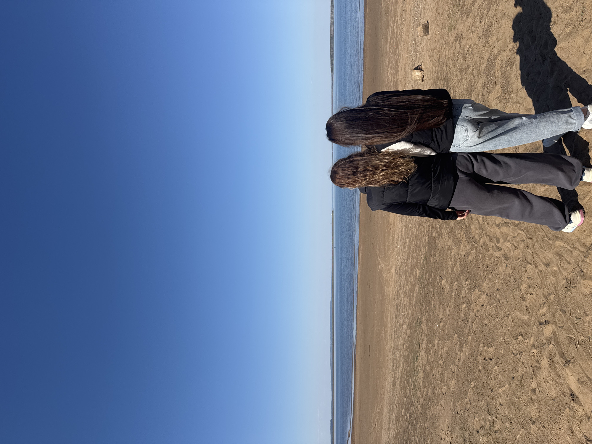 Two people are standing on a sandy beach next to a body of water. The person on the left has curly brown hair and is wearing dark sweatpants and a dark jacket. The person on the right has long, straight brown hair and is wearing light-colored jeans and a dark jacket. The sky is clear and blue. A shadow of one of the people is visible on the sand. The location is most likely the city of Chaikovsky, Russia, based on the user's request.