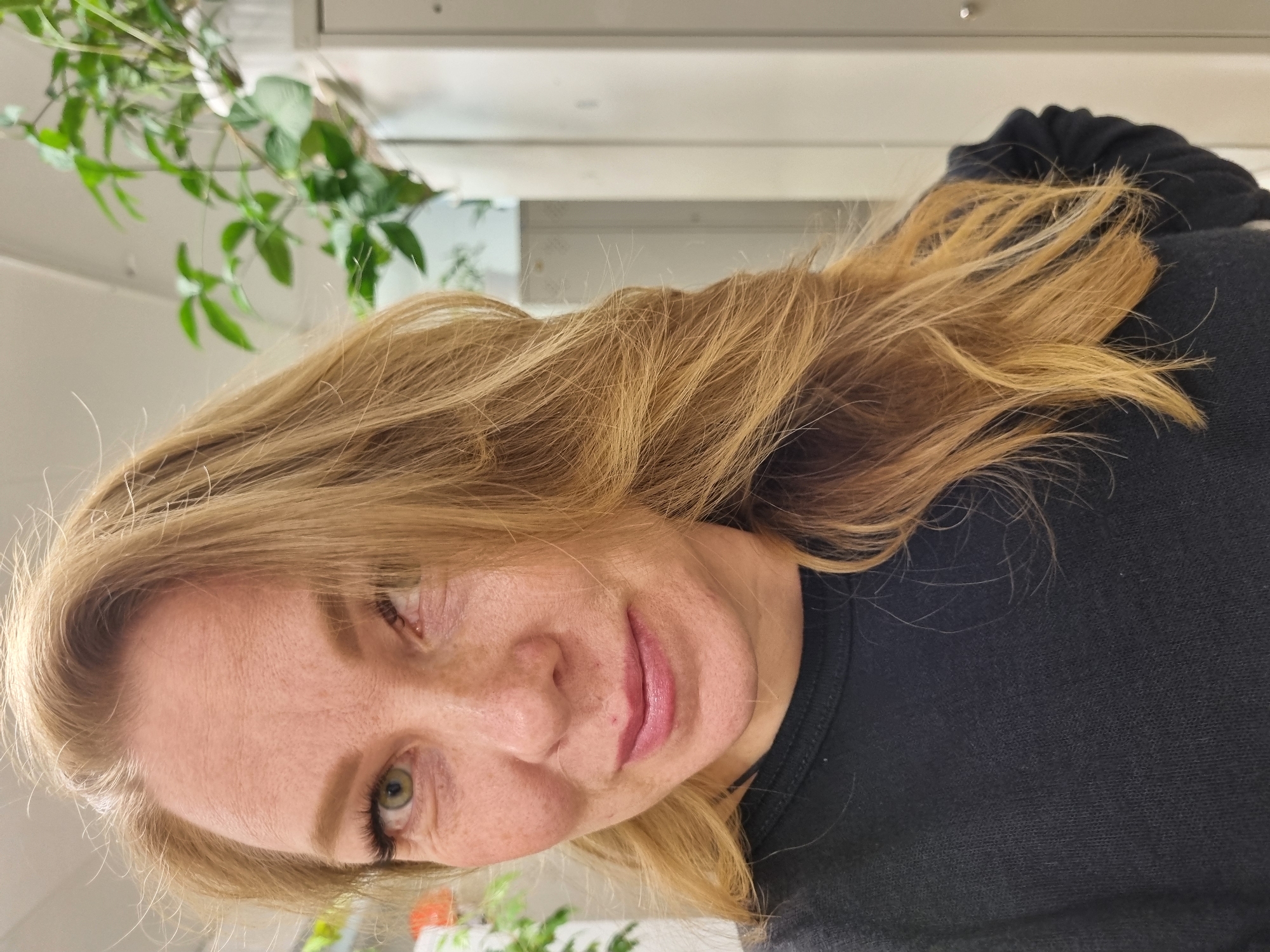 A woman with blonde hair, freckles, and green eyes is shown from the chest up, wearing a black shirt. The background is indistinct, showing a green leafy plant in the upper left and lower left, and a white wall behind. The image does not provide any information regarding location in Kemul, Russia.