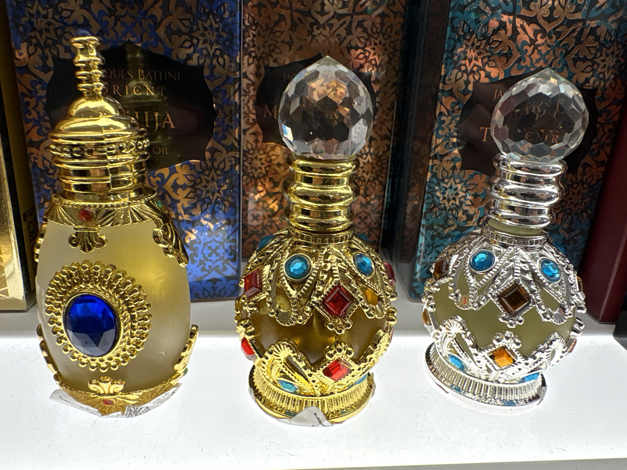 The image shows three ornate perfume bottles displayed on a white shelf. In the background, decorative boxes with intricate patterns are visible, suggesting they might be the packaging for the perfumes. The bottles themselves are highly detailed.
The leftmost bottle is made of a translucent, golden-hued material and is adorned with intricate gold filigree. A large, deep blue gem is prominently featured on the front of the bottle, set within a sunburst-like golden frame. The stopper is also gold and has a tiered, architectural design.
The middle bottle is made of a darker, amber-colored glass. It is extensively decorated with gold metalwork and encrusted with colorful jewels, including red squares, blue circles, and possibly amber or brown diamonds. The stopper is a large, faceted crystal, resembling a gemstone.
The rightmost bottle has a pale green, translucent body and is decorated with silver-colored metalwork that is also adorned with a variety of colorful gems. These include blue circles, brown diamonds, and orange or amber squares. Like the middle bottle, its stopper is a large, faceted crystal.
The lighting in the image is bright, highlighting the reflective surfaces of the bottles and their embellishments. The overall impression is one of luxury and elaborate craftsmanship. The scene appears to be a retail display, possibly in a perfume shop or department store. The location of the city and country are unknown.