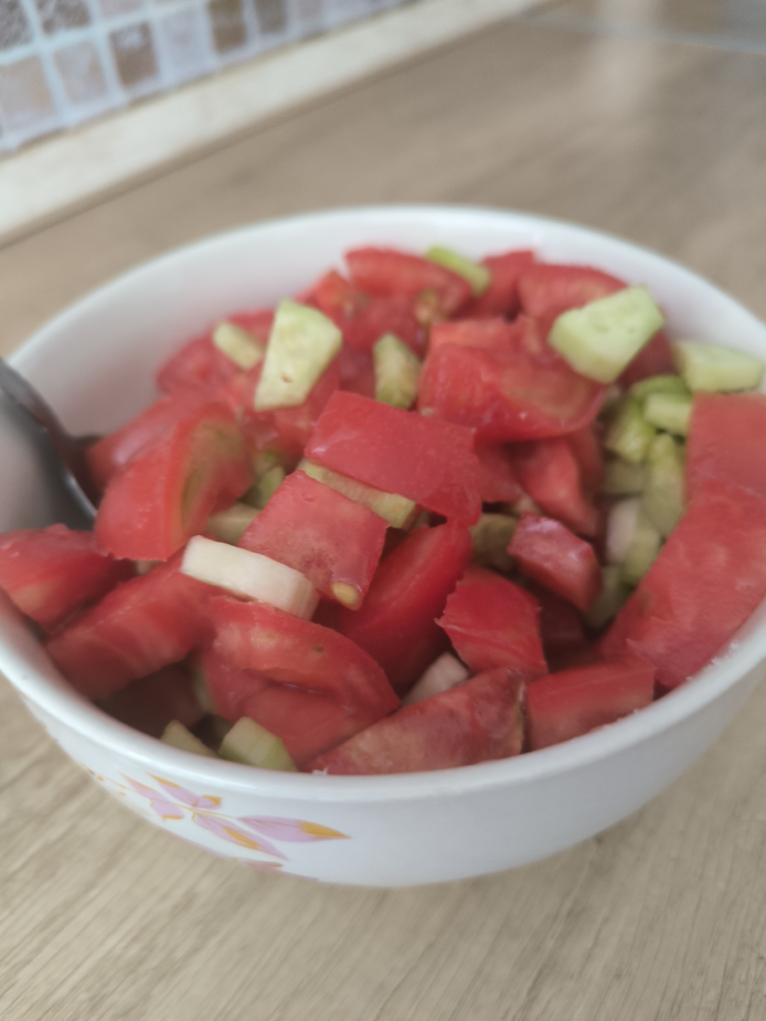 A close-up, eye-level shot shows a white bowl filled with chopped tomatoes and cucumbers. The bowl, which has a floral pattern on its exterior, sits on a light brown wooden surface. A metal spoon is visible to the left, partially submerged in the salad. The background is blurred, showing a beige and brown tiled wall. There is no specific indication of the location within Chișinău, Moldova, as the image focuses solely on the food and its immediate surroundings.