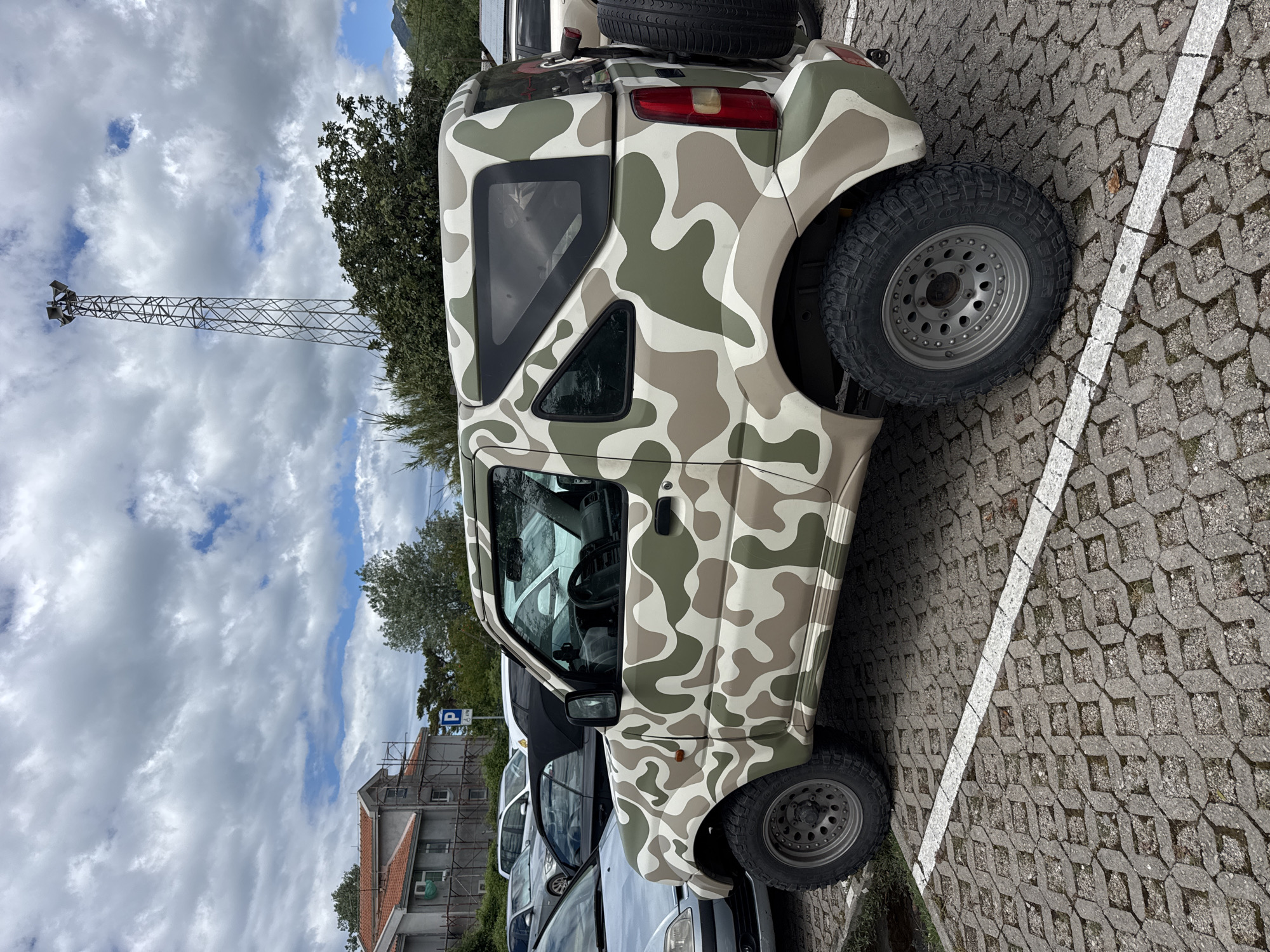 The image depicts a camouflage-painted Suzuki Jimny parked in a parking lot. The parking lot has cobblestone-like pavement marked with white parking lines. The sky above is cloudy. A tall, metal communication tower is visible in the background. Other cars are parked nearby. The location is in the city of Bar, Montenegro, as indicated by the presence of signs and buildings characteristic of the area. The image shows a still, outdoor scene.