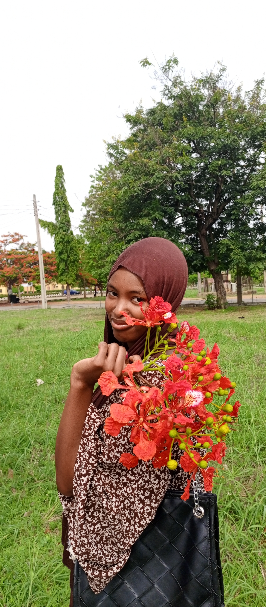 A young woman wearing a brown hijab and a patterned brown and white floral dress stands in a grassy area, holding a bouquet of vibrant red flowers with green buds. She is slightly smiling and looking at the camera. A black quilted handbag hangs from her arm. The background includes green trees, a lawn, and a building with a red tree nearby. Utility poles are also visible. This image was likely taken in Keffi, Nigeria.