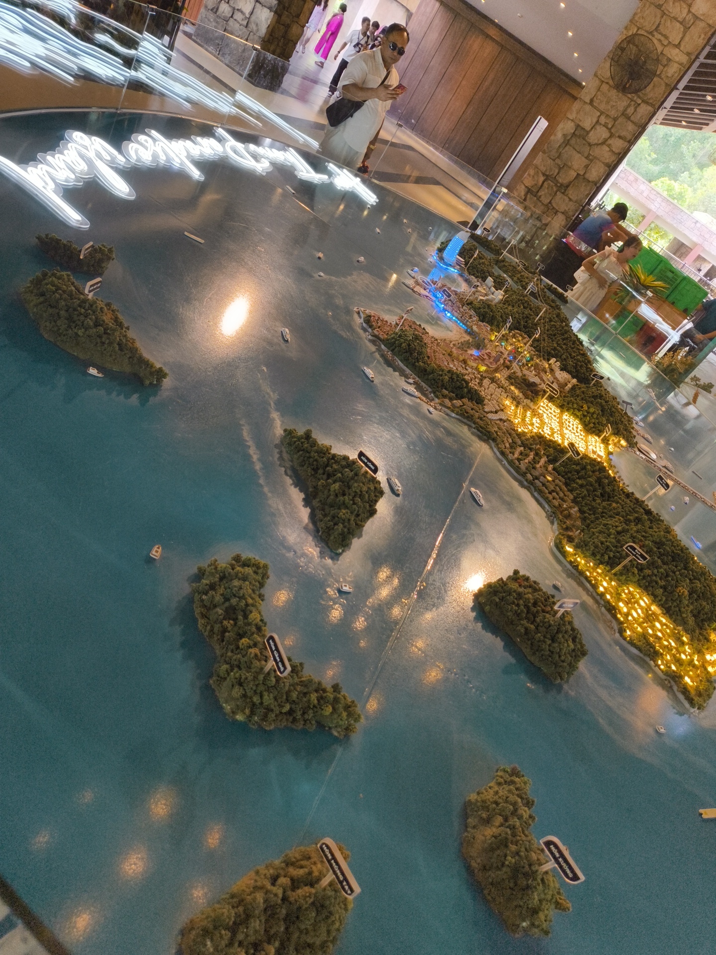 A diorama of islands and a city is displayed on a table with a blue, water-like surface. The diorama features a densely built-up city illuminated by yellow lights, surrounded by green, tree-covered islands. Several miniature boats are placed on the water. In the background, people are visible, some looking at the diorama. The environment appears to be indoors, possibly a museum or exhibition space, with stone walls and a wooden ceiling. The city depicted is likely part of Phú Quốc, Vietnam, given the context of the model and signage visible on some islands.