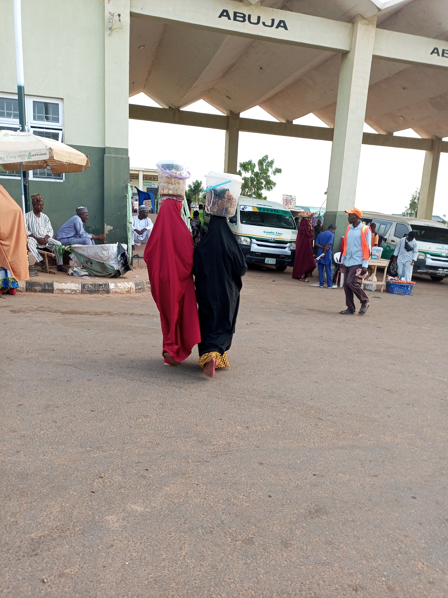 The image depicts an outdoor scene at a bus station in Gombe, Nigeria. The location appears to be a waiting area with a covered structure. In the foreground, two women are walking away from the camera, each carrying a large bucket of goods on their heads. They are both wearing long, flowing robes that cover them from head to toe. The woman on the left is wearing a bright red robe, and the woman on the right is wearing a black robe. Their feet are visible, and they are both wearing sandals.
In the mid-ground, several people are visible. To the left, three men are sitting on a bench under a large umbrella. Two of them are wearing traditional caps. Behind them, a man in a blue outfit is standing near a white bus with "TOYOTA" and "GOMBE LINE" written on it. To the right of the bus, a man wearing an orange cap and a reflective vest is walking. He is looking towards the left. Another man, wearing a light blue outfit, is standing near a small wooden table with a blue crate on it. Further in the background, more people and vehicles are visible, suggesting a busy transportation hub.
The environment is paved with asphalt, and there are some trees and vegetation visible in the background. The overall lighting suggests it is daytime. The text "ABUJA" is visible on the overhead structure, indicating this might be a terminal for buses traveling to or from Abuja. The presence of the "GOMBE LINE" on the bus further supports the location being in Gombe, Nigeria.