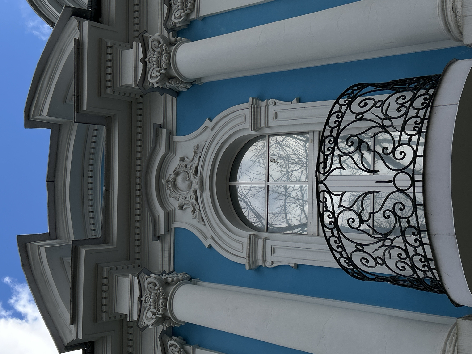 The image captures a section of the exterior architecture of the Smolny Cathedral located in Saint Petersburg, Russia. The architecture features a blue wall with white columns, ornate carvings, and a rounded window with a black decorative iron balcony. The reflection in the window suggests bare tree branches, possibly indicating a winter or late autumn scene. A portion of the blue sky with clouds is visible above the architectural structure.