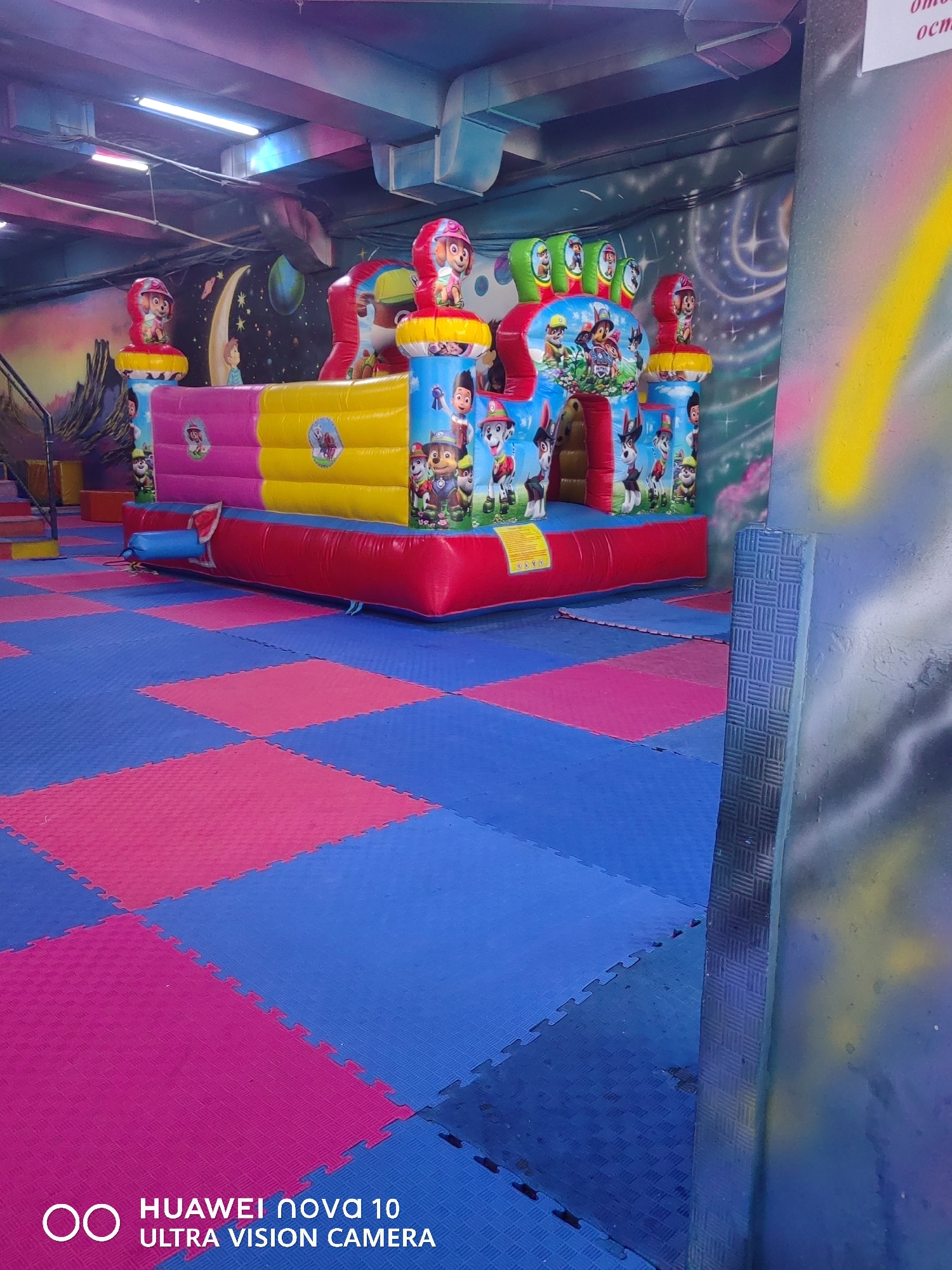 The image shows an indoor children's play area in Özgön, Kyrgyzstan. A large, inflatable bouncy castle decorated with characters from the Paw Patrol franchise is the central focus. The bouncy castle has multiple colorful sections, including pink, yellow, and red, and features images of the cartoon dogs. It is situated on a floor covered with interlocking foam mats, alternating in blue and pink. In the background, a mural depicting space and celestial bodies adorns the wall. The ceiling is visible with fluorescent lighting fixtures. To the right, a textured pillar with graffiti, including a yellow stripe and black and grey spray paint, is partially visible. The overall environment appears to be a commercial entertainment venue for children.