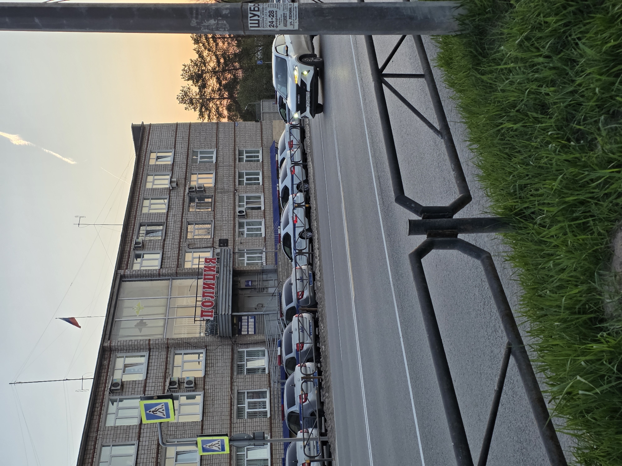 The image depicts a Russian police station located in the city of Chaikovsky, Russia. The station is a multi-story brick building with multiple windows and air conditioning units. The word "ПОЛИЦИЯ" (Police) is displayed prominently in red lettering above the entrance. Several police cars are parked in a row along the street in front of the station. A white SUV is visible at the front of the line of parked vehicles. A pedestrian crossing sign is also visible near the building. A grey metal railing runs along the edge of the pavement with grass growing on the far side. A streetlight or pole is positioned on the left side of the image. A sign advertising furs (ШУБЫ), sheepskin coats (ДУБЛЕНКИ) and Yak fur boots (УНТЫ ИЗ ЯК) during an event from February 24-28 on Lenin Street (ул. ЛЕНИНА) is present on the light pole. The sky is visible in the background, suggesting either sunset or sunrise.
